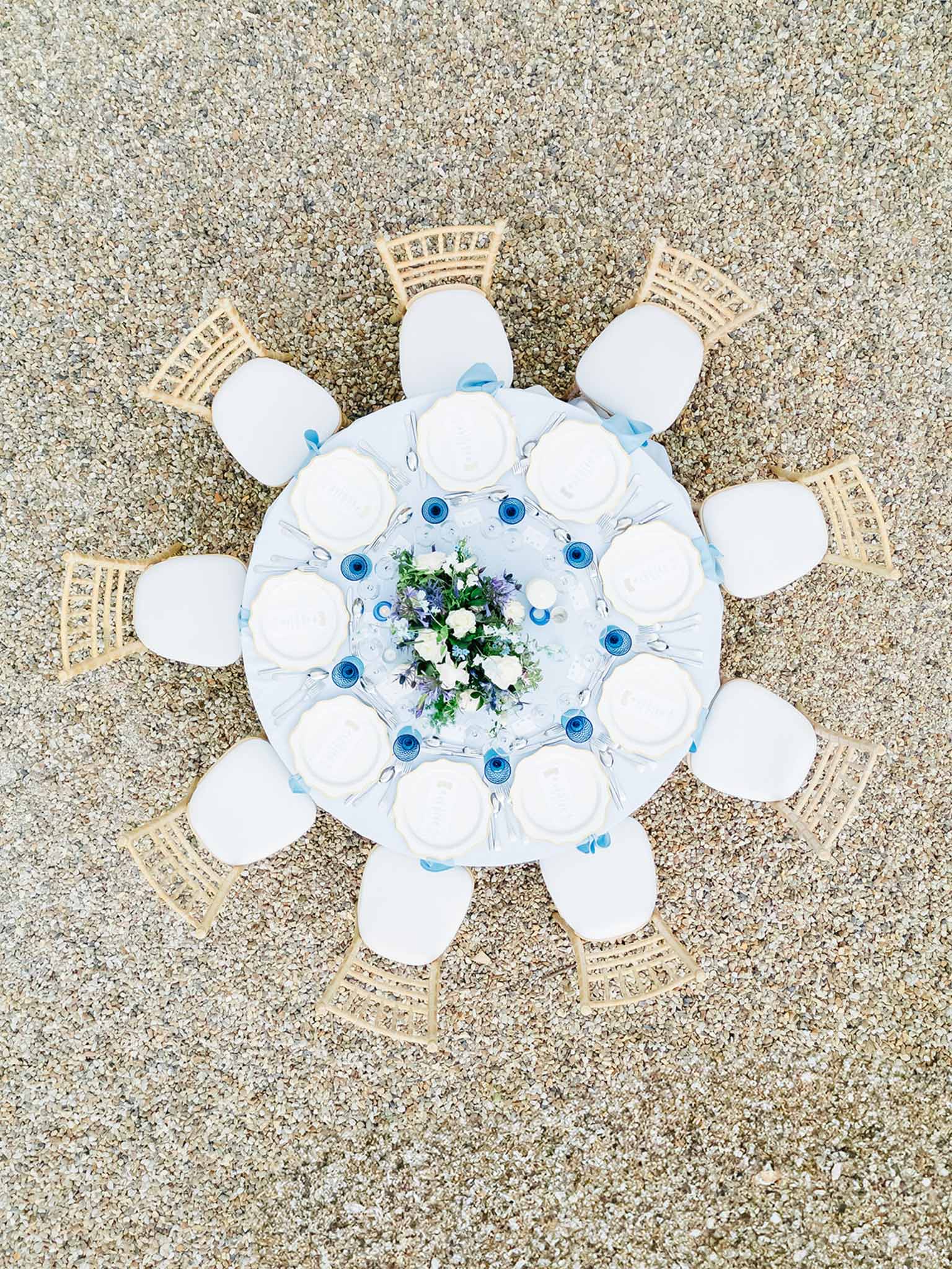 Aerial view of round table set on sand with blue linens, rattan chairs, and blue delphinium centerpiece