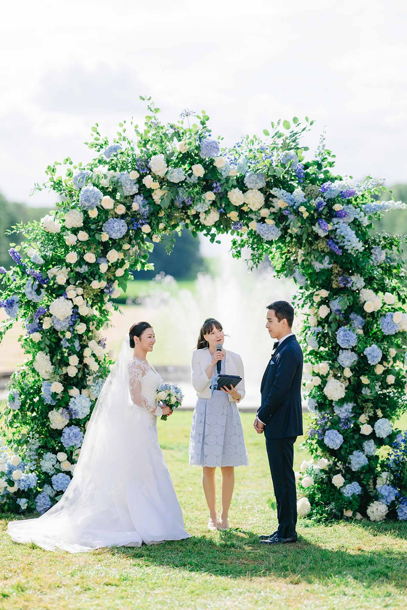 Bride and groom under hydrangea arch in ivory and blue with officiant during outdoor garden ceremony