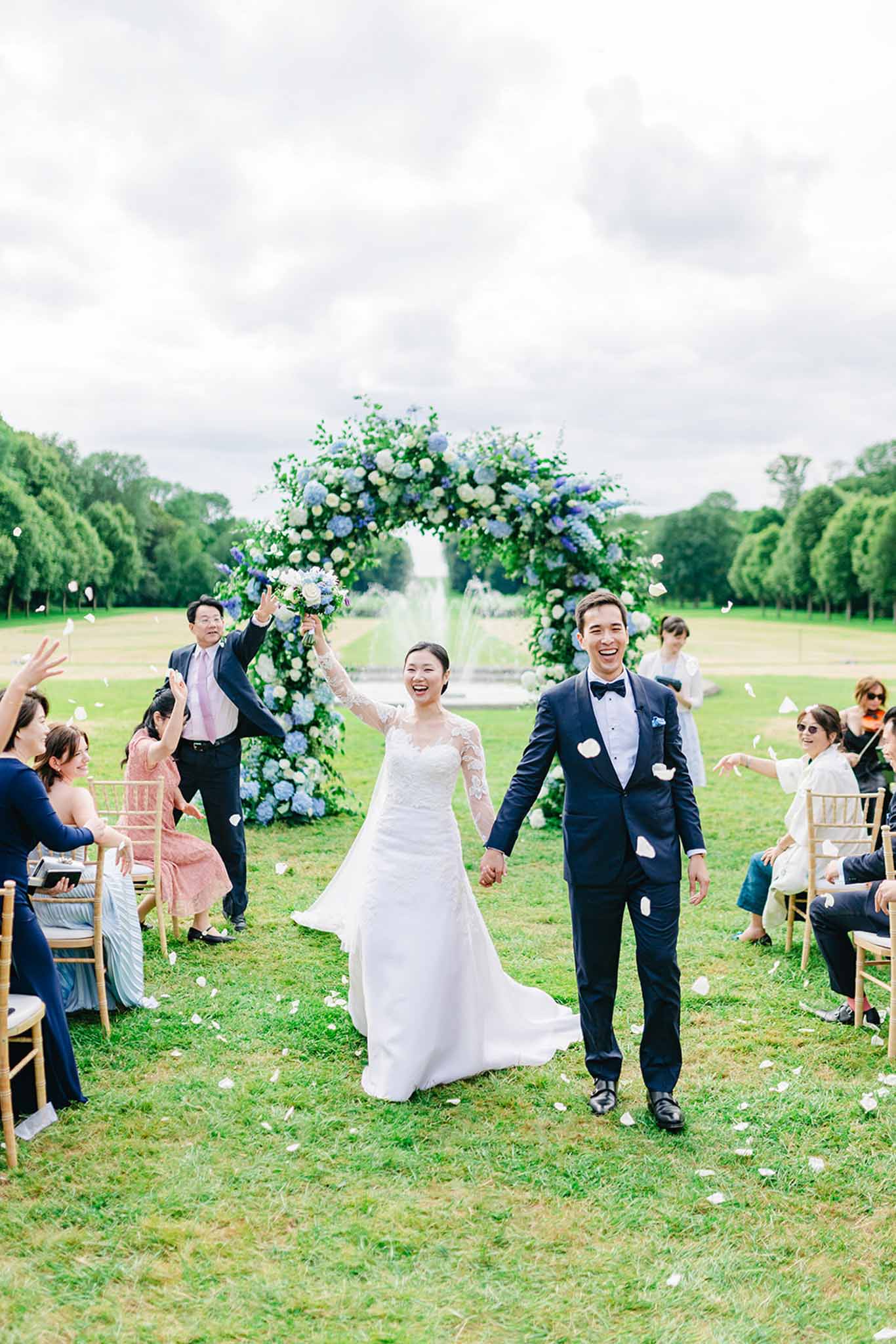 Bride and groom recessional through white hydrangea arch with guests throwing petals at formal French canal garden