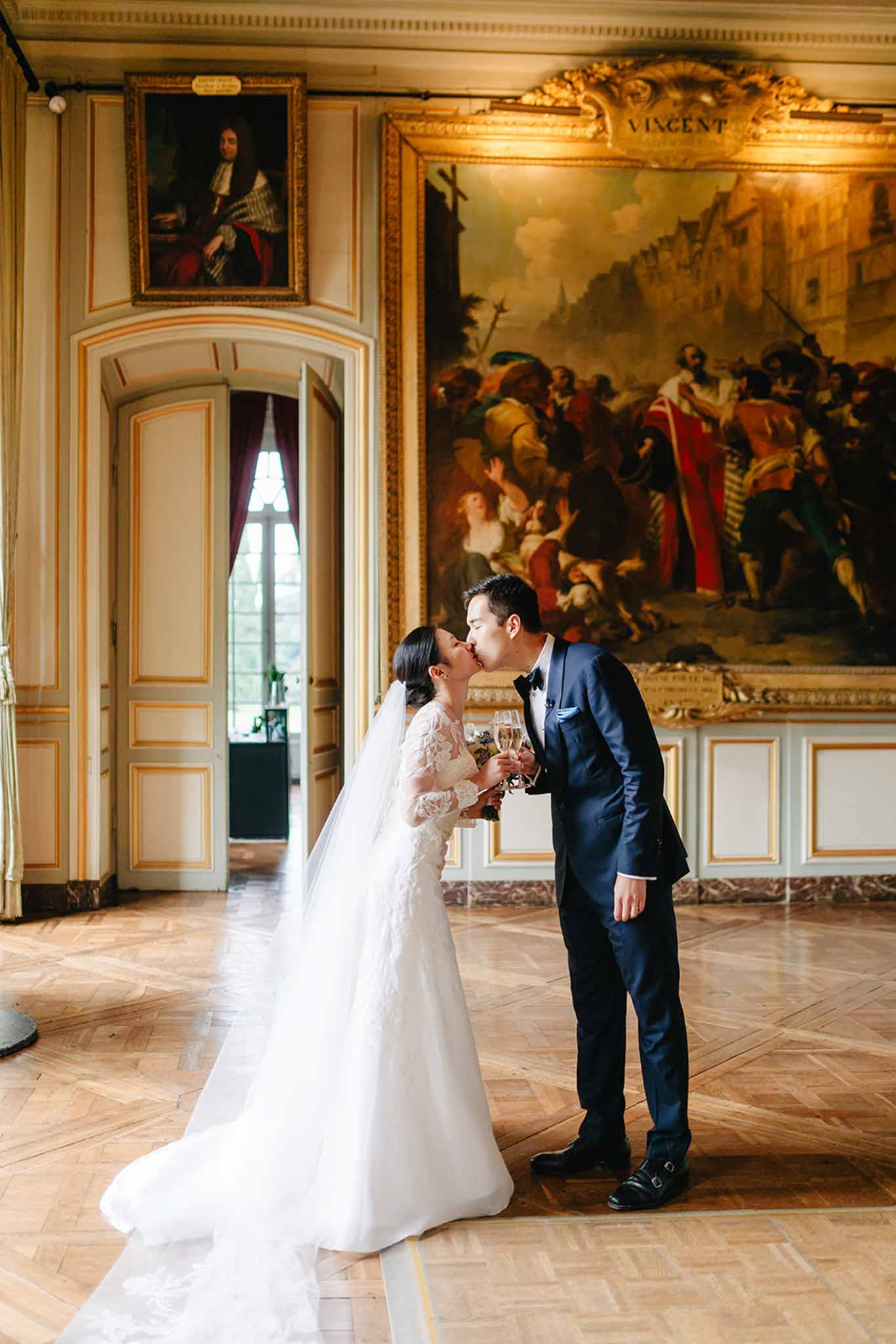 Bride and groom kissing in ornate classical ballroom, bride in ivory lace gown holding white bouquet, groom in navy suit