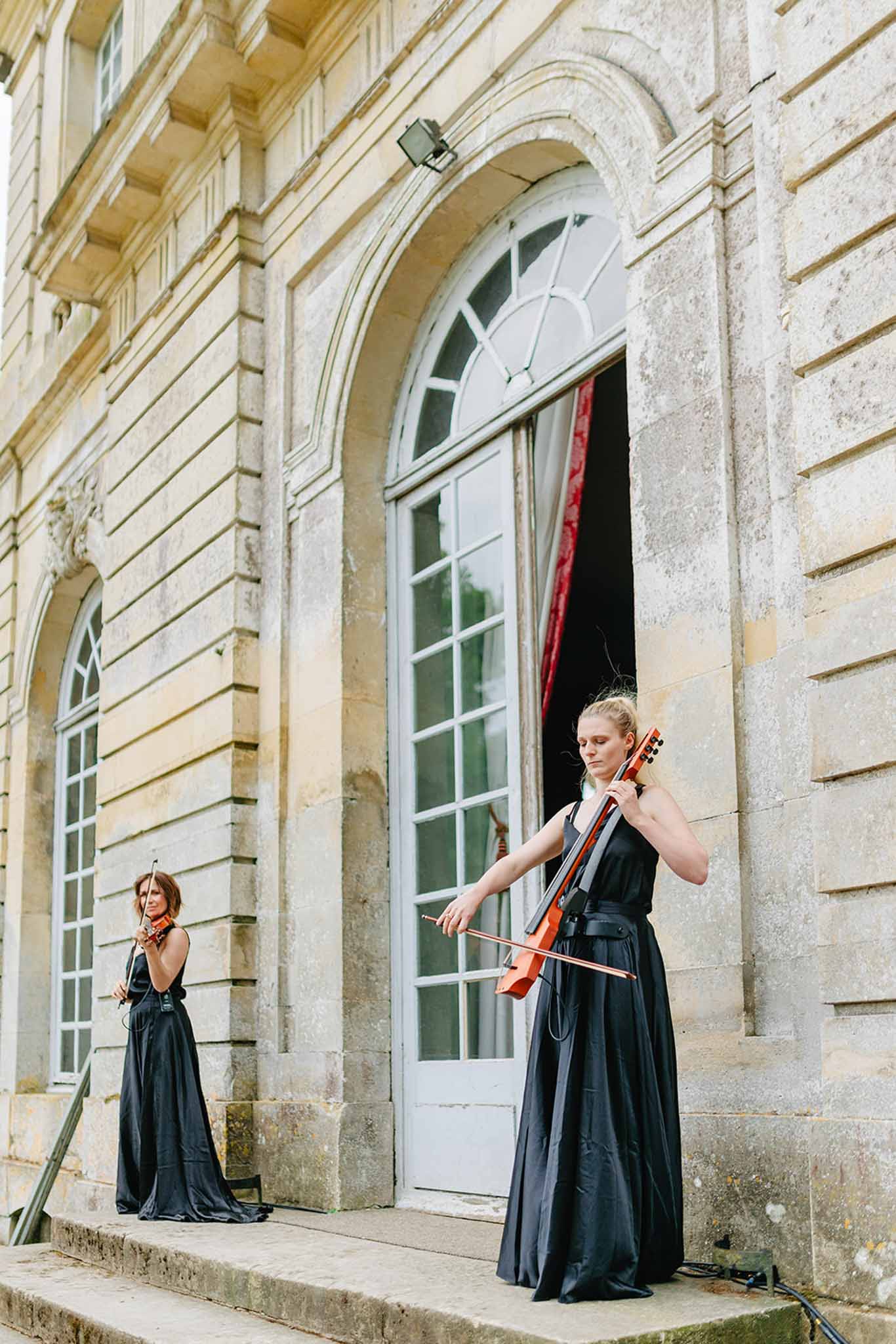 Two musicians in black dresses outside classical stone building, cellist in foreground, violinist beside arched window