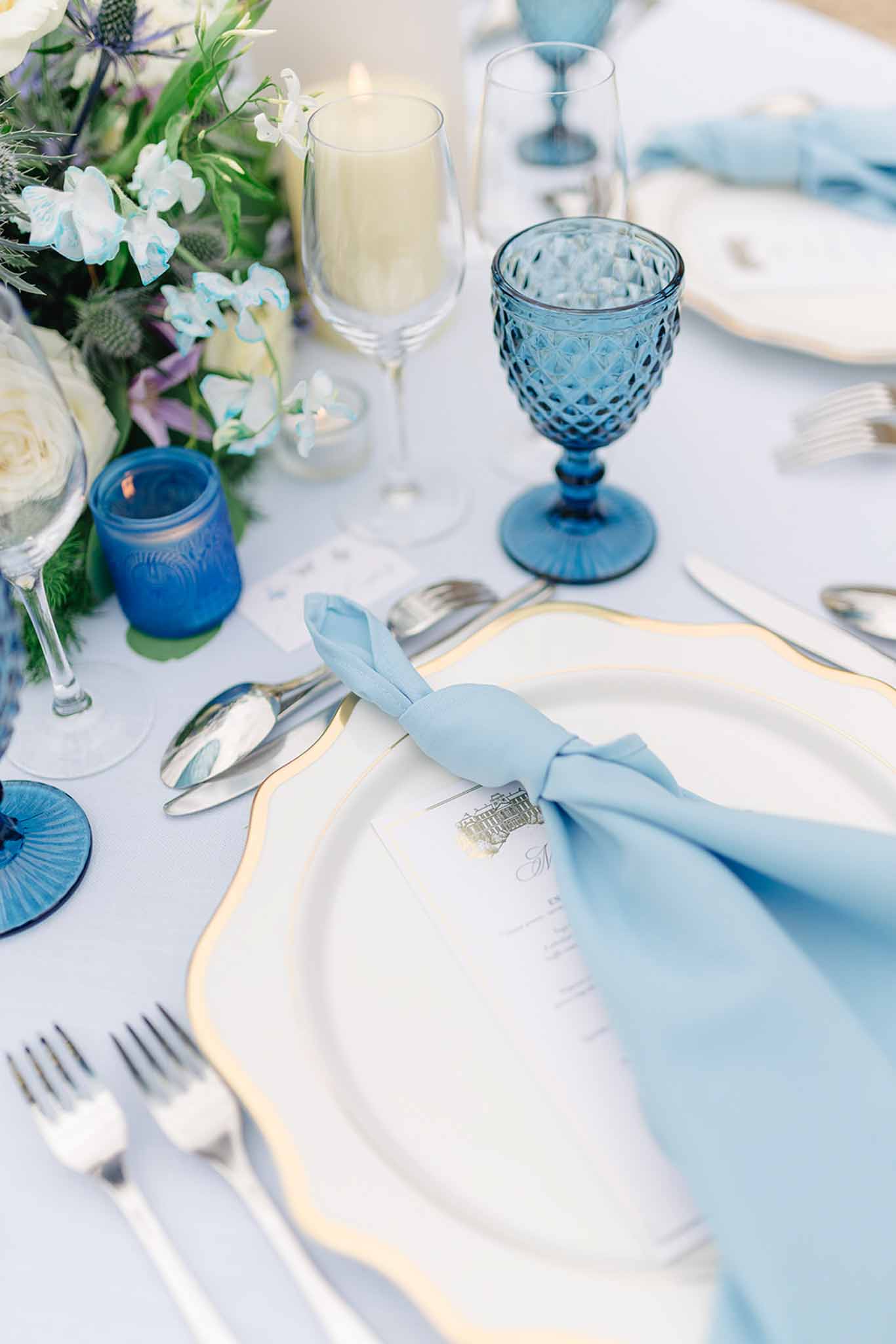 Reception place setting with blue goblet, gold-rimmed white plates, light blue napkin and eryngium floral centerpiece