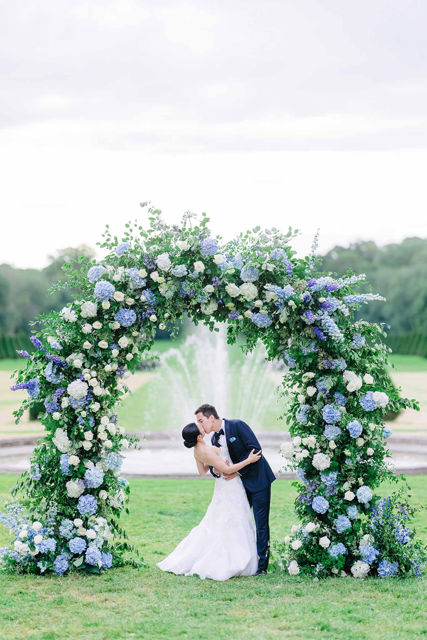 Bride and groom kissing under blue hydrangea and white rose ceremony arch on lawn
