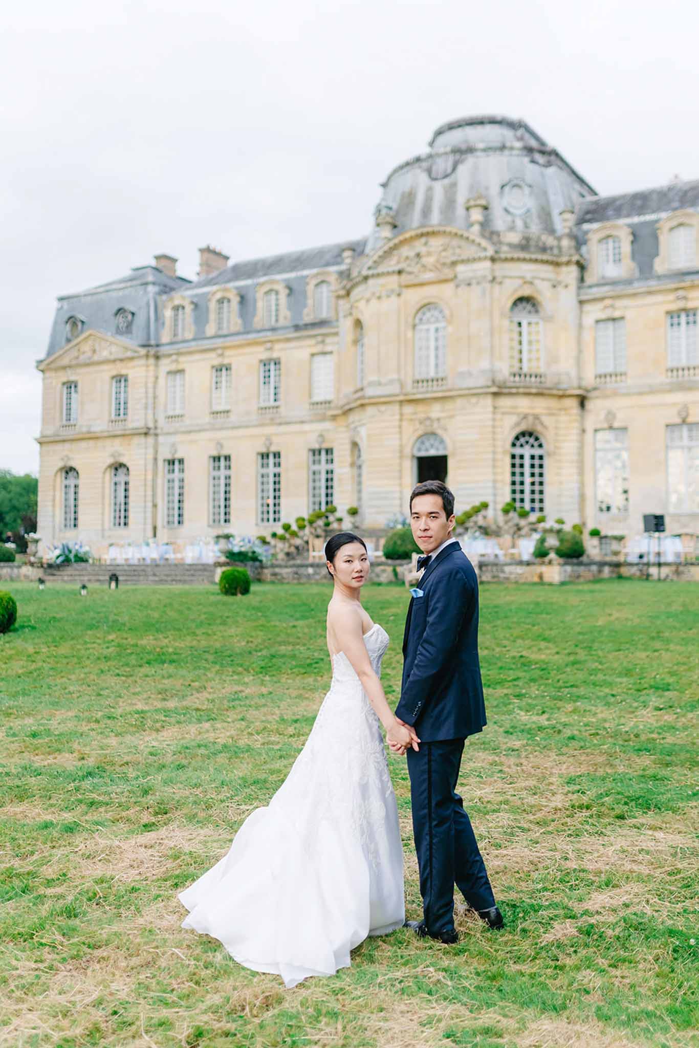 Bride and groom posing on lawn before grand classical chateau with mansard roofs at Champlatreux