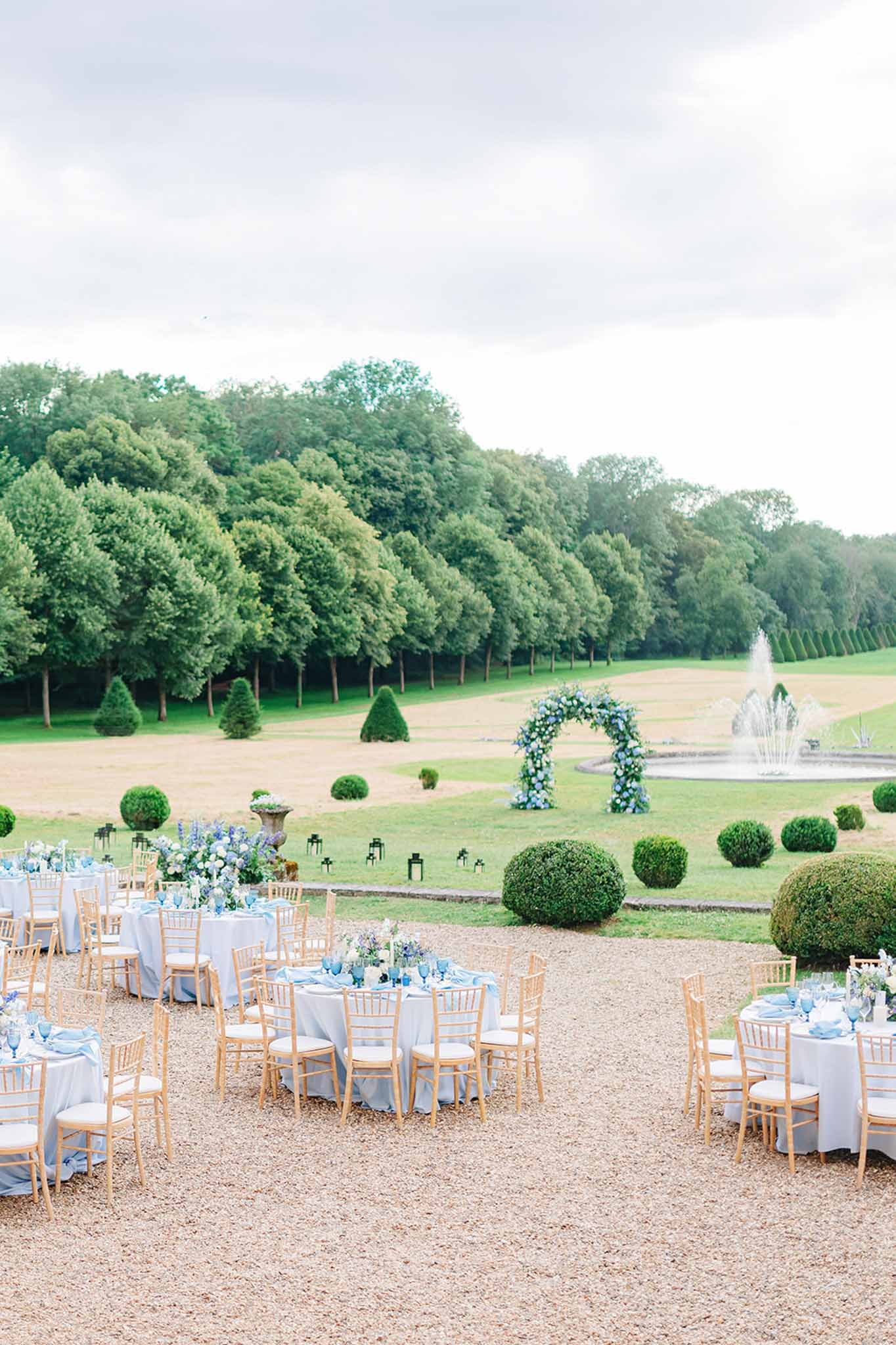 Outdoor reception with round tables in pale blue linens and ceremonial arch in formal French garden