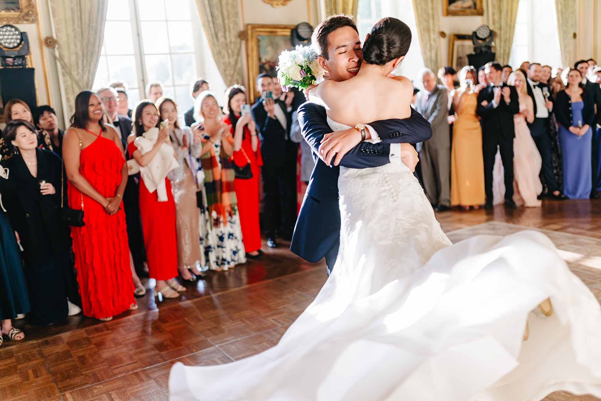 Couple sharing first dance in grand ballroom with classical curtains and guests in formal attire watching