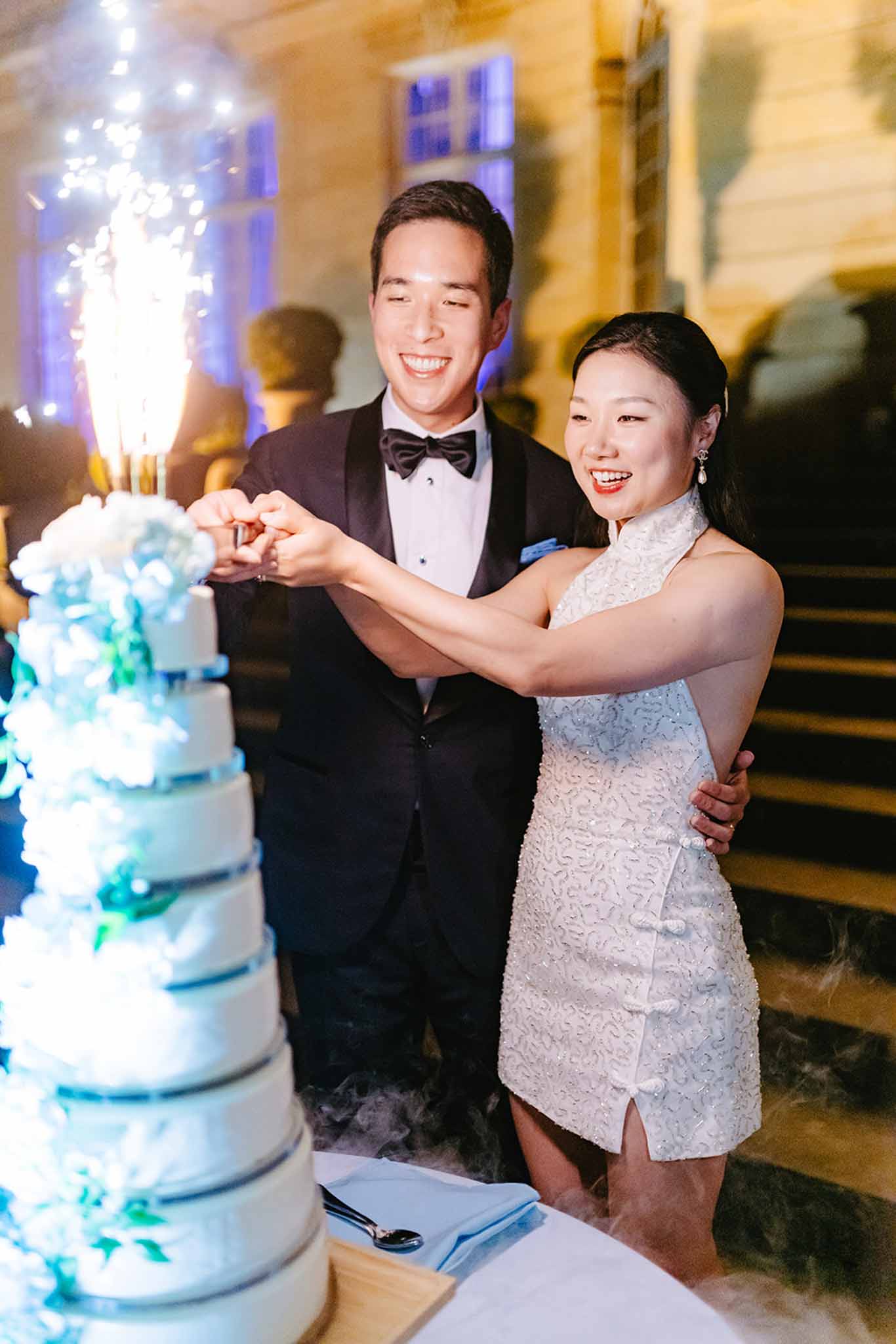 Bride and groom cutting three-tier white cake with turquoise floral piping and sparkler topper at evening reception