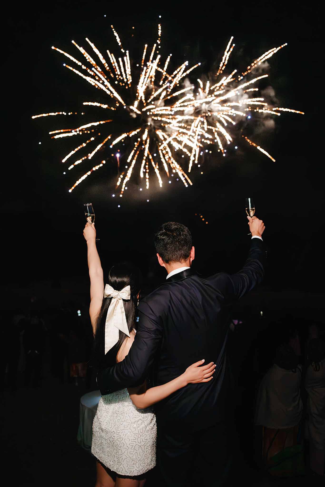 Bride and groom raising champagne flutes toward gold fireworks in the night sky