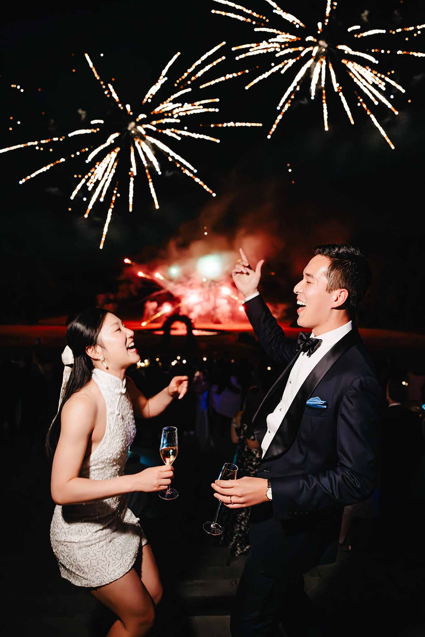 Bride in short ivory dress and groom in black tuxedo laugh as white and gold fireworks burst overhead at reception