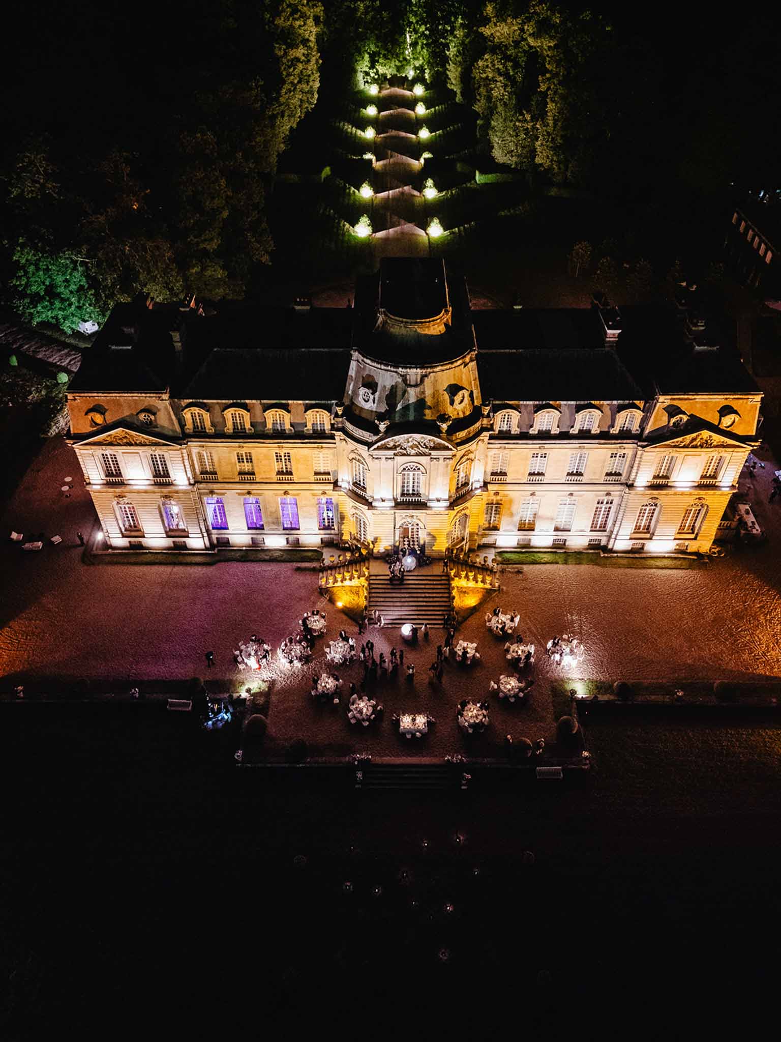 Aerial view of nighttime courtyard reception at chateau with 150-200 guests dining among candlelit tables and uplighting