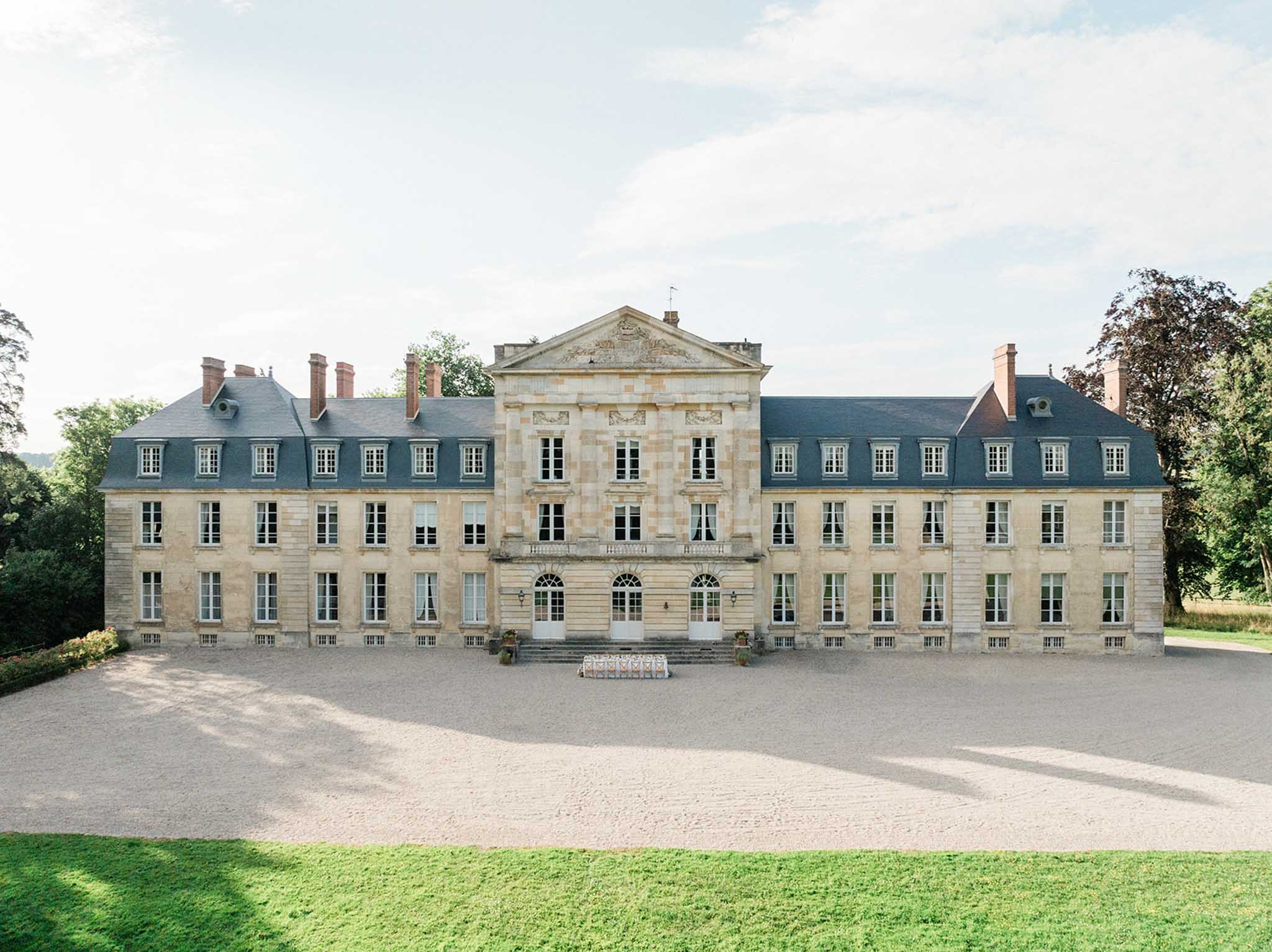 Wide symmetrical view of an 18th-century French chateau with mansard roofs, gravel forecourt, and manicured lawn