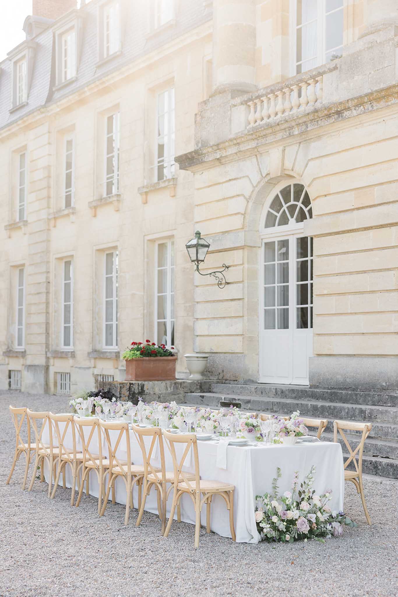 Long reception table set on gravel forecourt of 18th-century stone château with blush and white floral arrangements