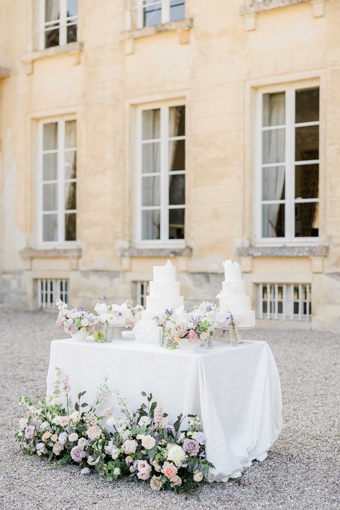 Two white tiered wedding cakes surrounded by blush and lavender roses on a table in a stone courtyard
