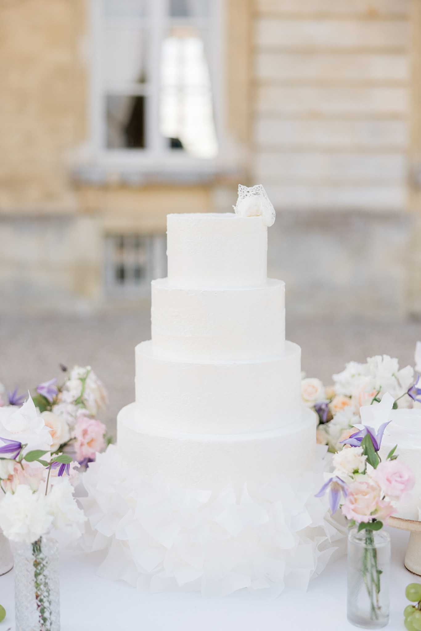 Five-tiered white wedding cake with lace topper flanked by pastel floral arrangements in outdoor courtyard