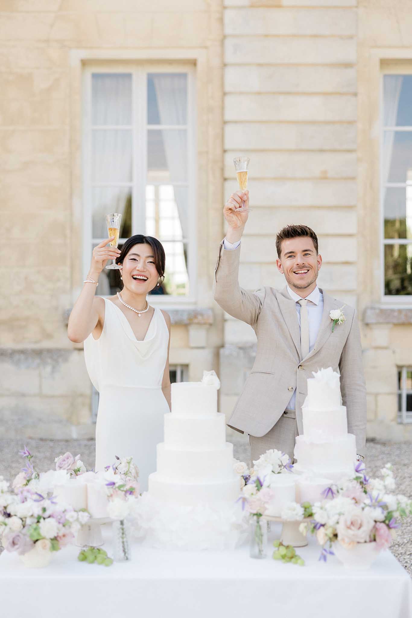 Bride and groom toasting beside multi-tiered white wedding cake flanked by pink and purple floral arrangements