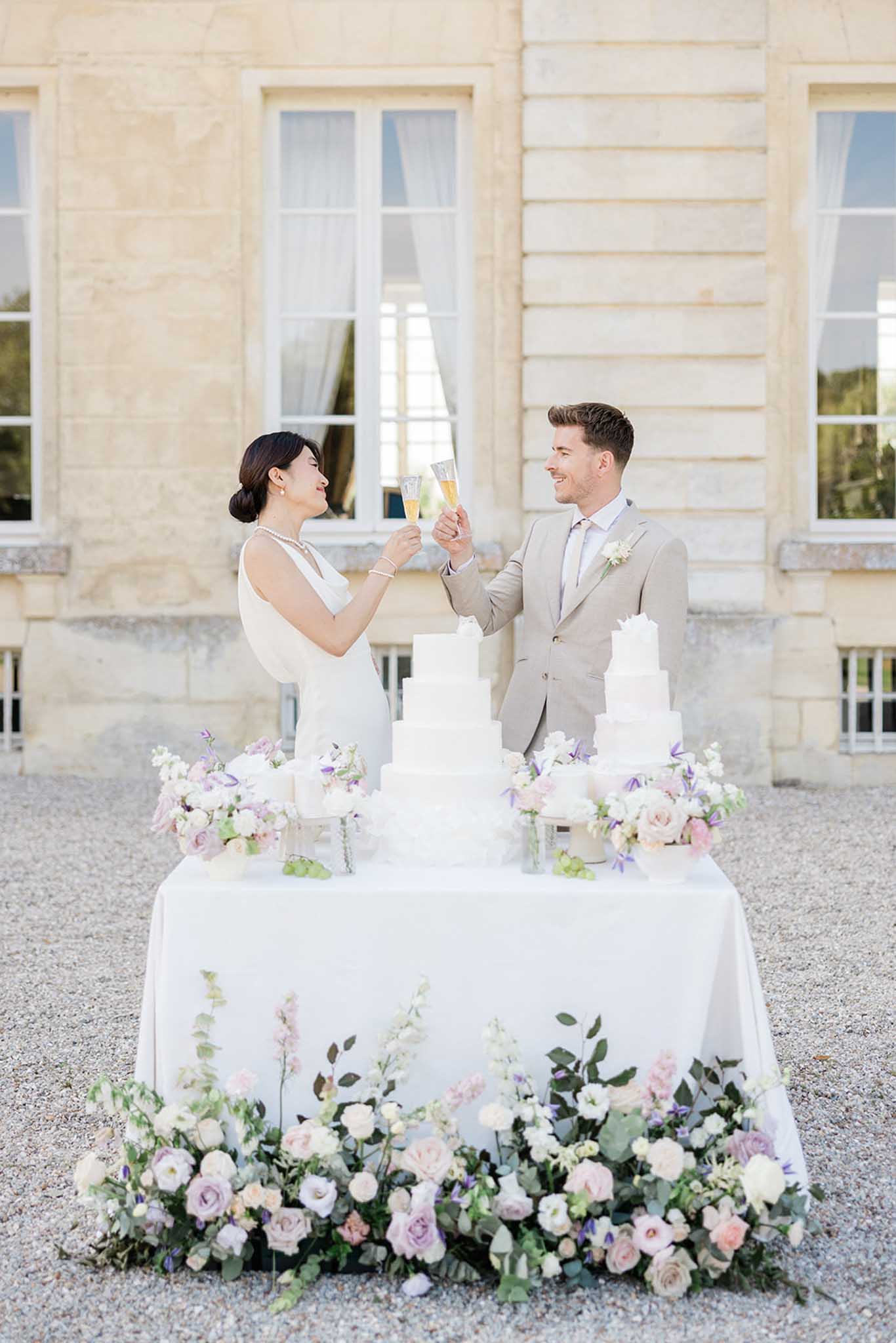 Bride and groom toasting by multi-tiered white wedding cake on courtyard table flanked by blush roses and lavender floral arrangements
