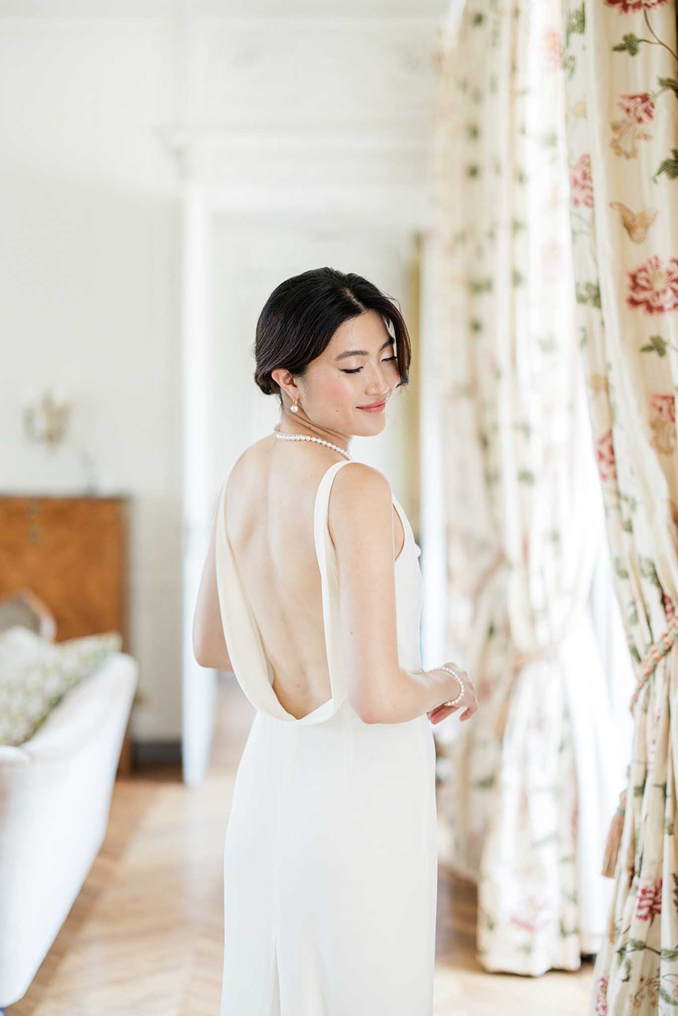 Bride in backless ivory gown with pearl necklace, dark hair in soft updo, looking down in a floral-curtained bedroom during preparation.