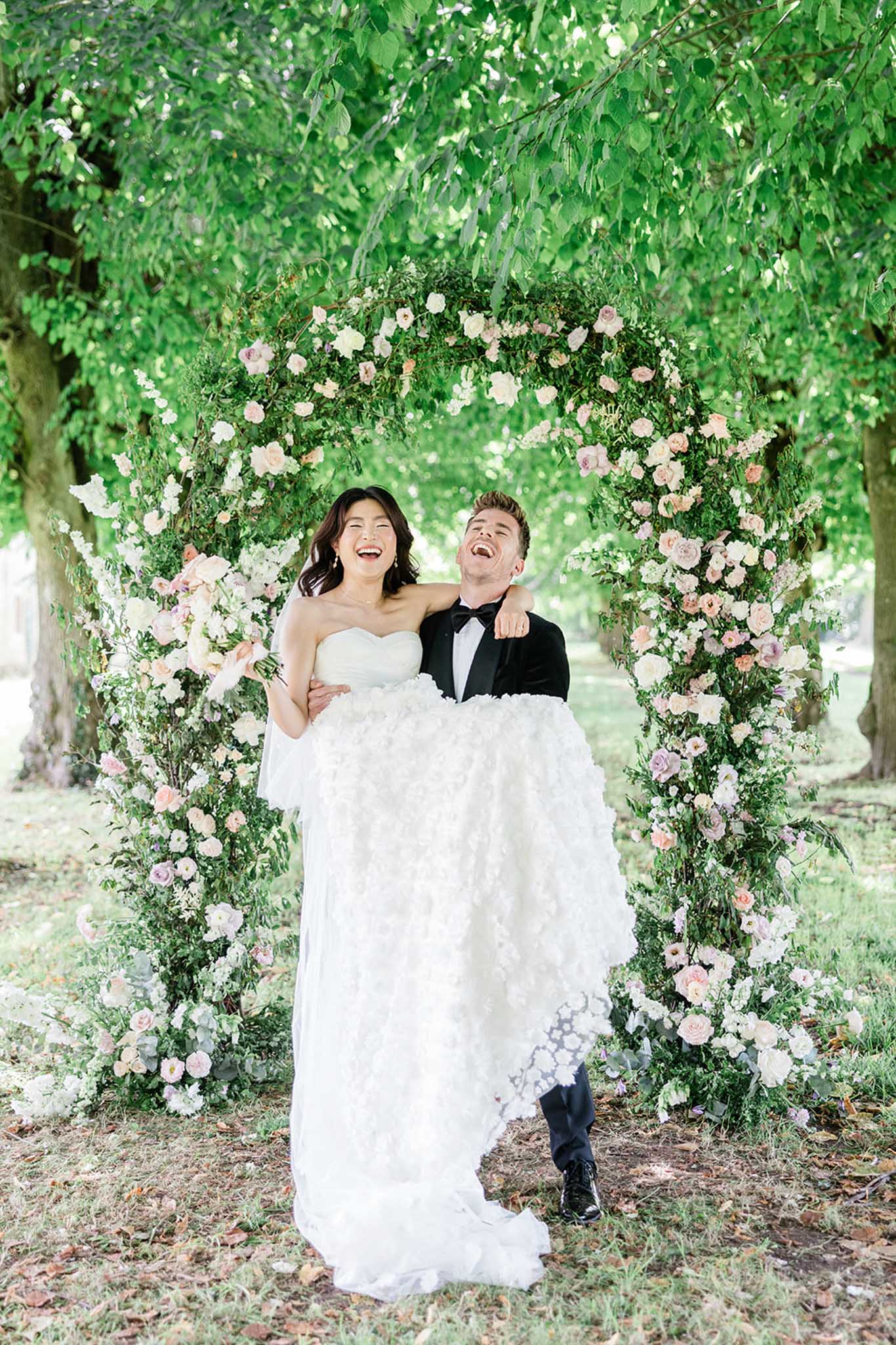 Groom carrying bride beneath lush pink and white rose floral arch in garden, both smiling upward