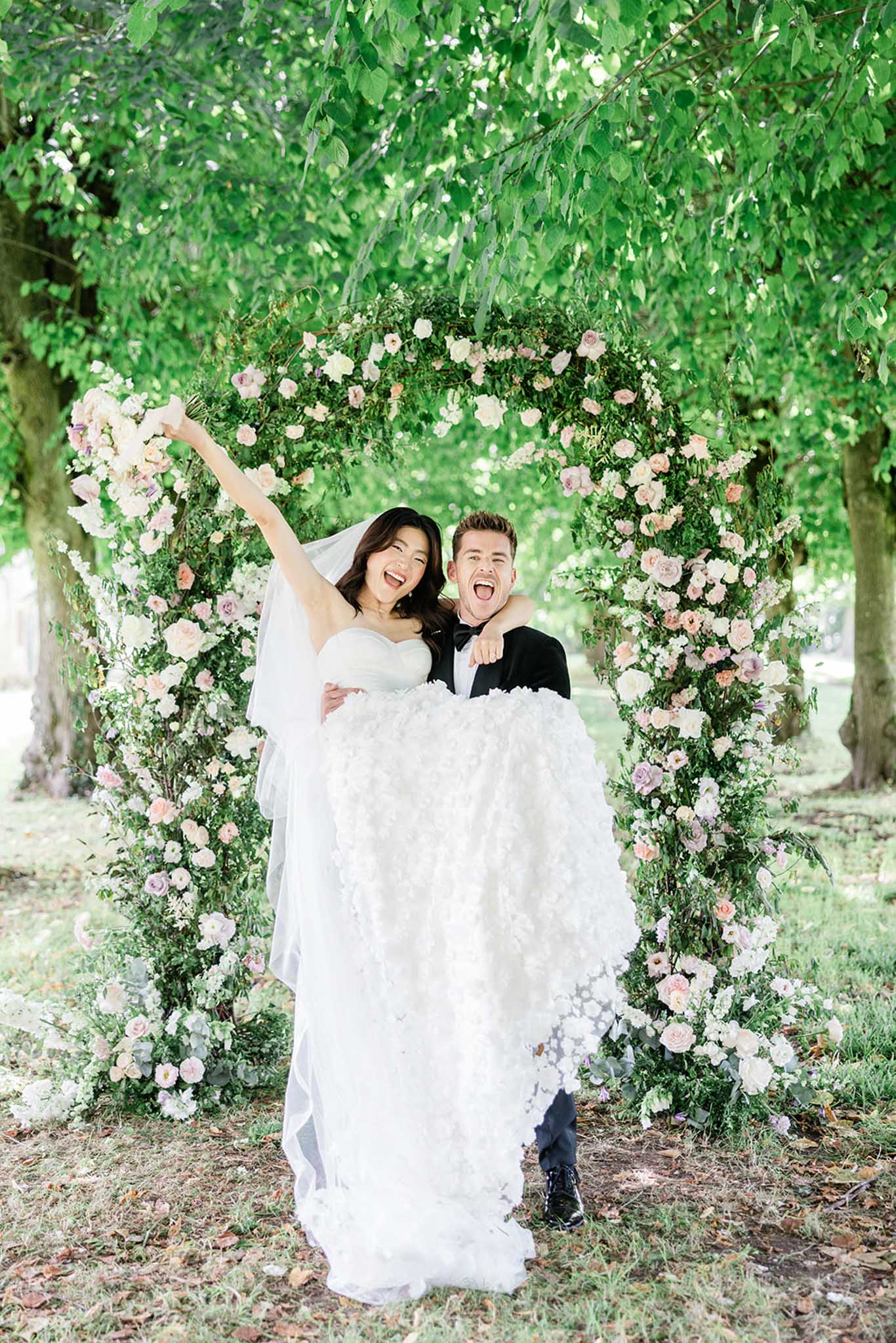 Groom carrying bride beneath blush pink and ivory rose floral arch in garden