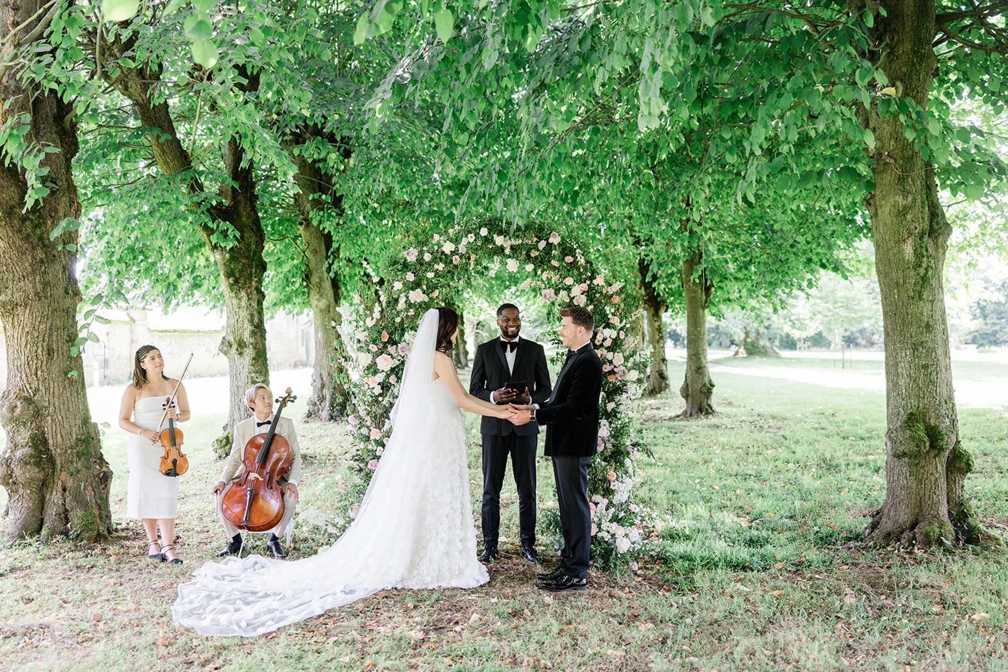 Outdoor ceremony beneath tree-lined alley with floral arch and live musicians at Chateau de Courtomer