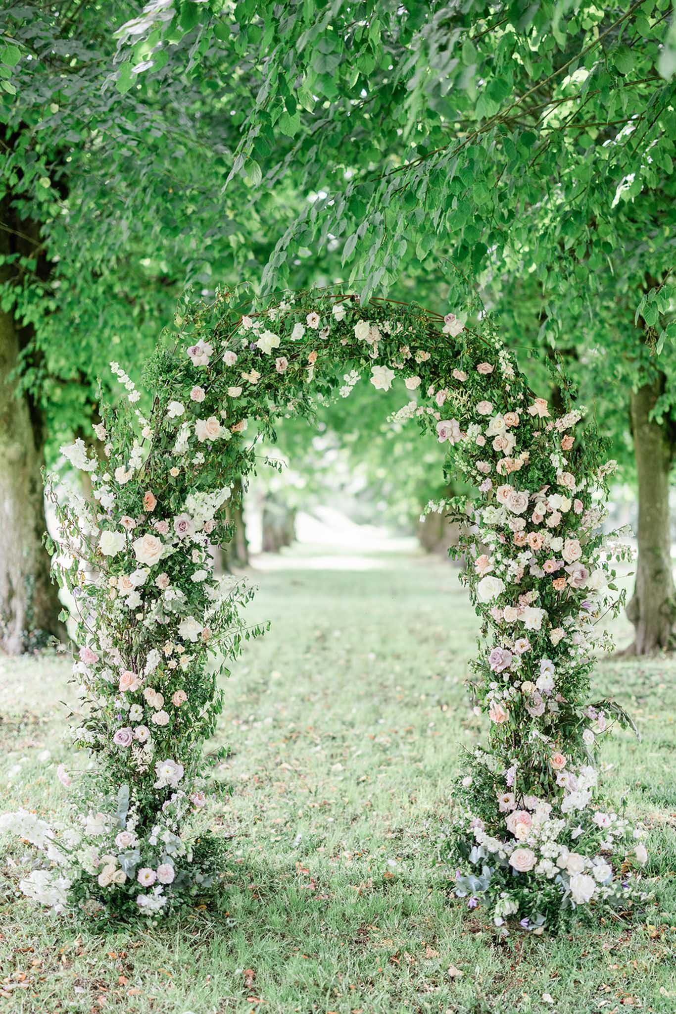 Floral ceremony arch with blush roses and greenery in tree-lined garden pathway