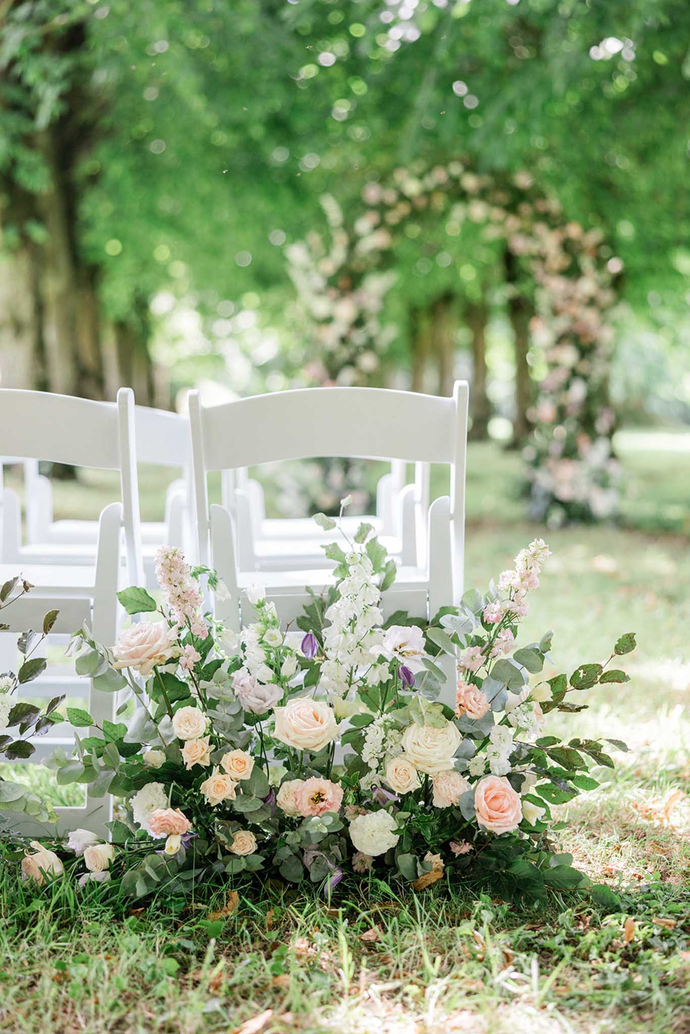 Lush floral arrangement of blush and cream roses with eucalyptus at base of white Chiavari chairs along garden ceremony aisle