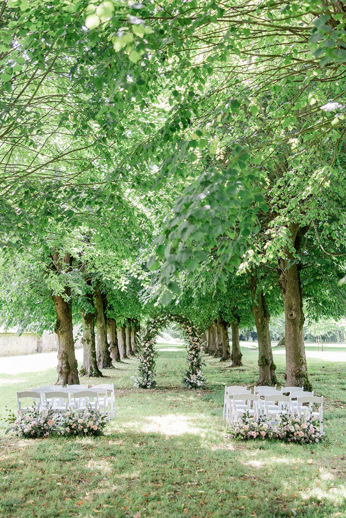 Tree-lined ceremony aisle with white folding chairs and vine-wrapped floral arbors at a chateau garden
