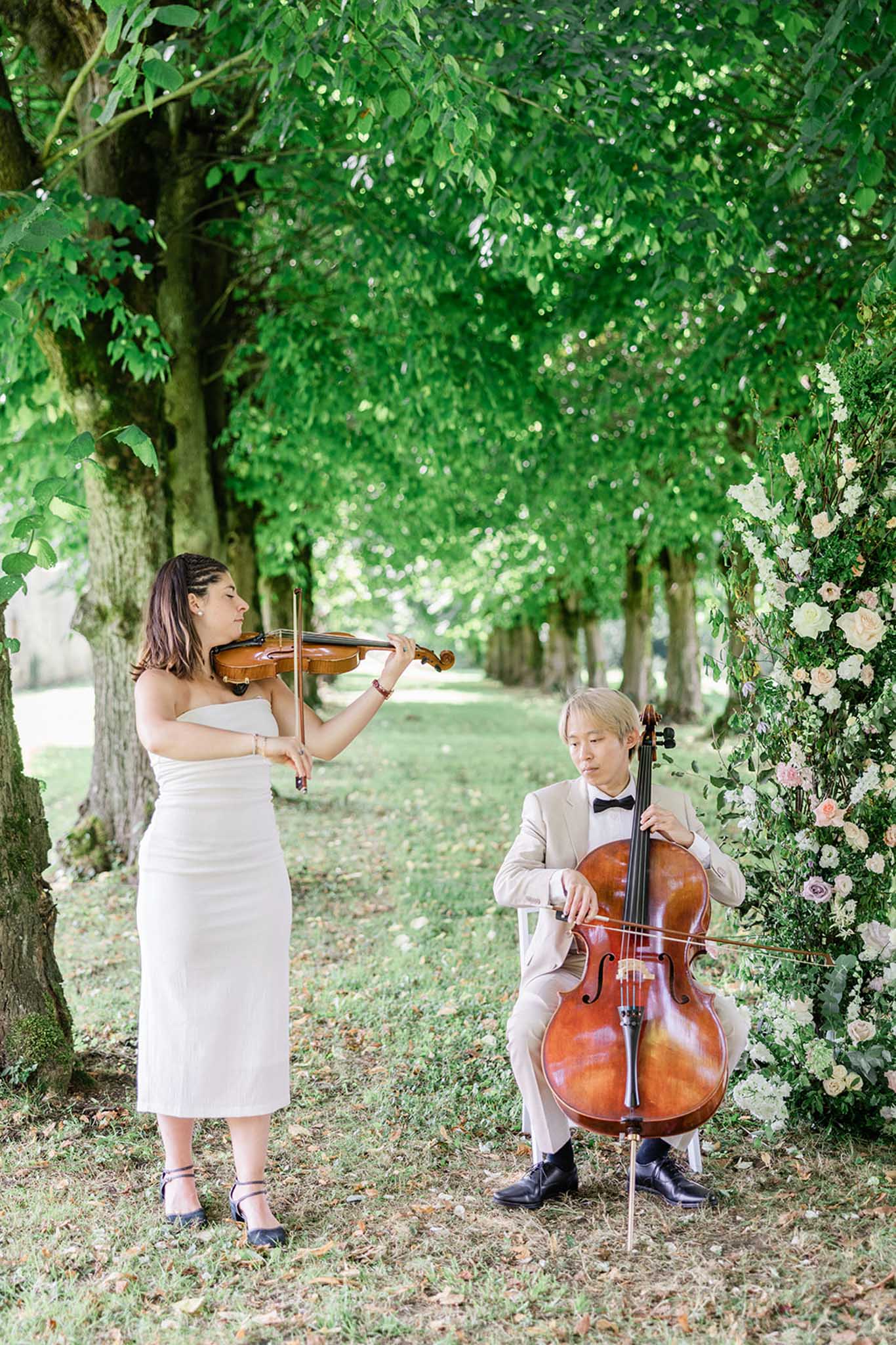 Violinist and cellist performing beneath tree-lined alley during outdoor wedding ceremony with rose trellis behind