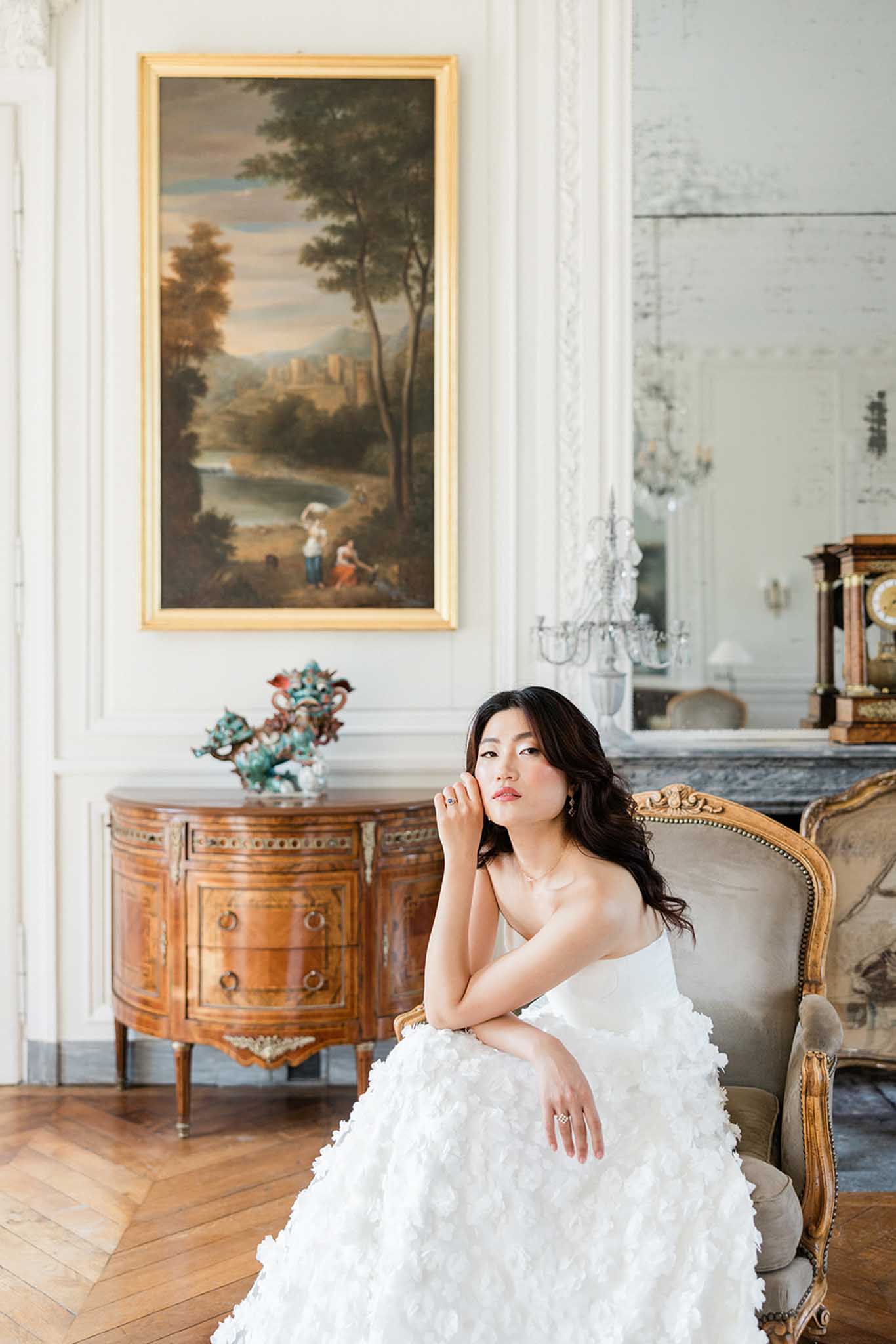 Bride in textured ivory gown seated in gold-leaf armchair in classical interior with landscape painting above
