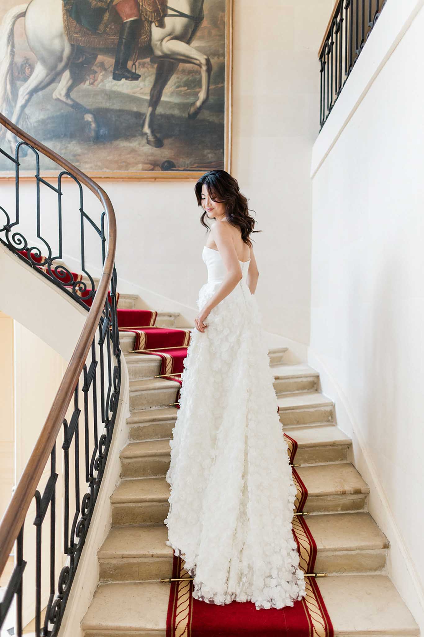 Bride in ivory ruffled gown descends red-carpeted stone staircase past equestrian painting in chateau interior