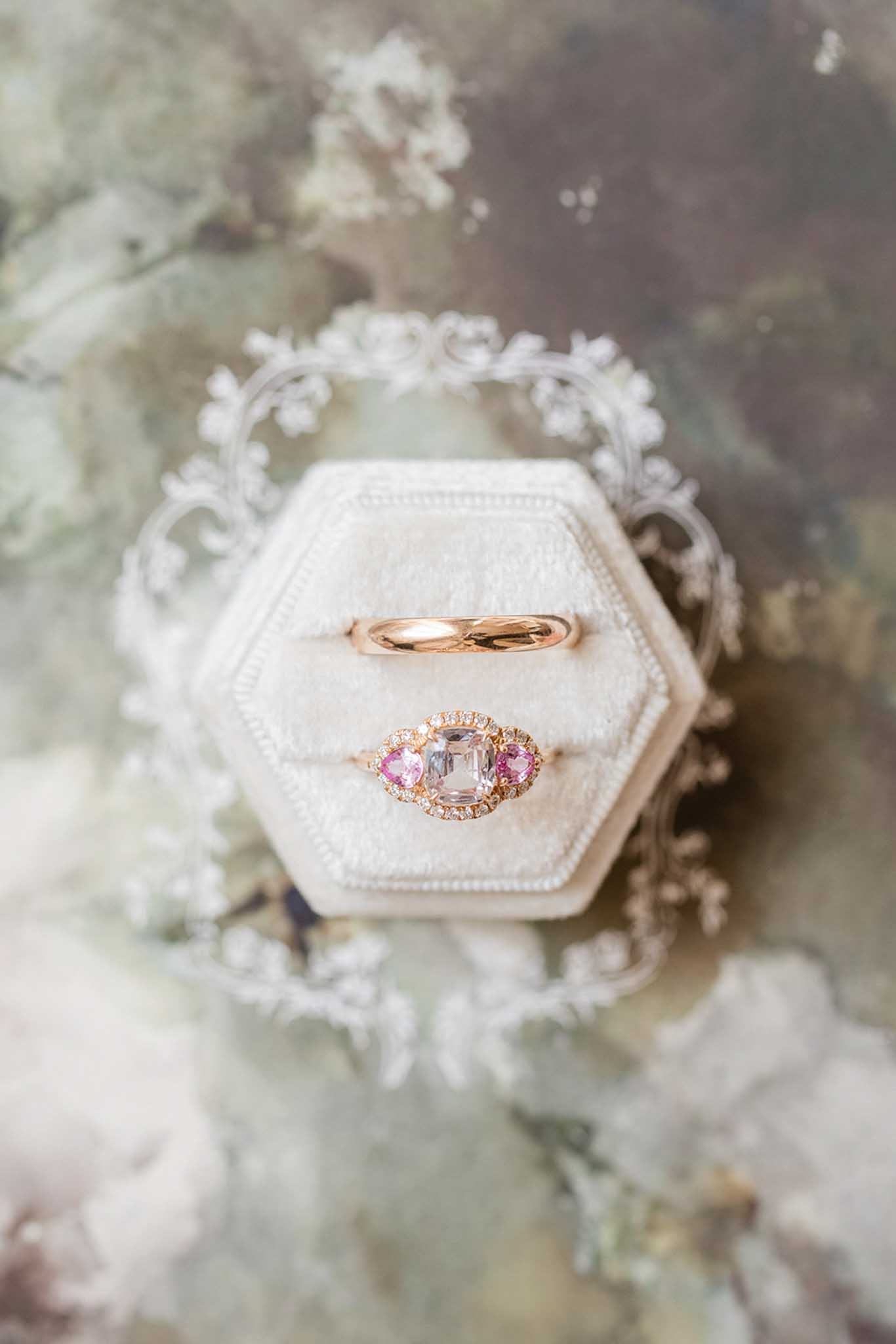 Rose gold ring with pink morganite and tourmalines beside curved rose gold band in white velvet box with flowers