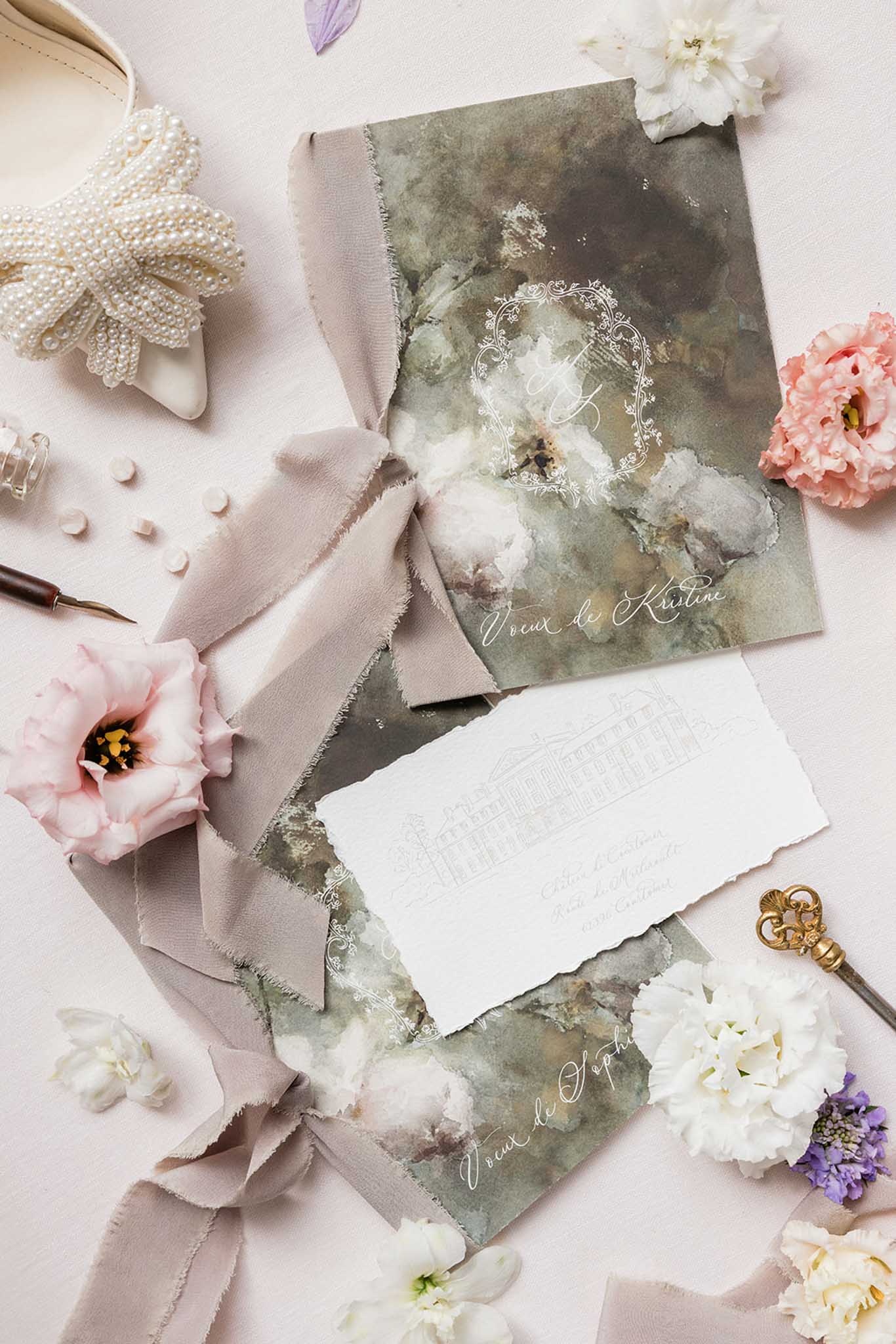 Flat lay of wedding stationery with sage green watercolor invitation, coral peonies, pearl beads, and dusty rose ribbon