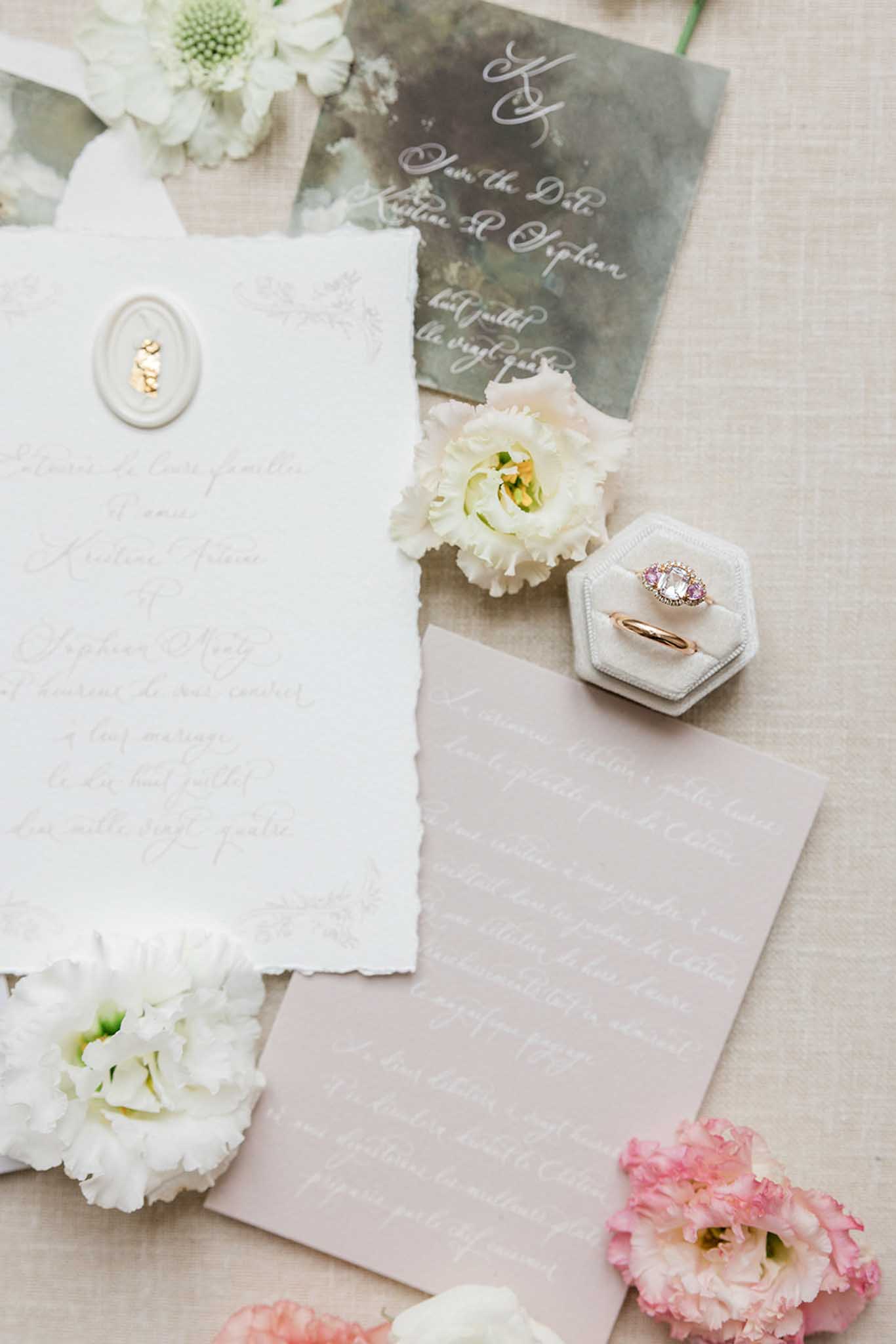 Wedding stationery flat lay with ivory invitation, sage vellum card, engagement ring, and white peony and dahlia blooms