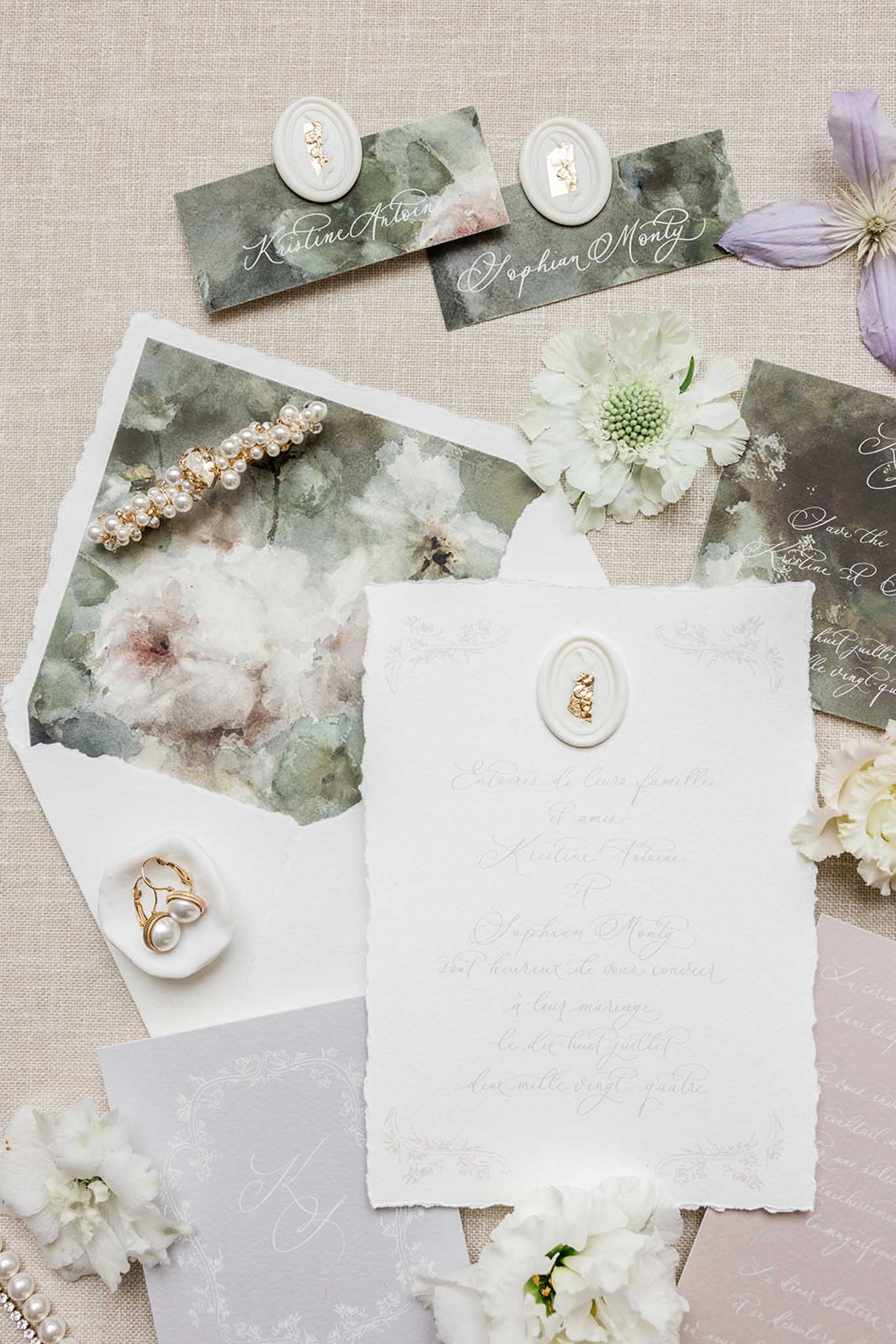 Flat lay of wedding stationery, place cards, wax seals, pearl hair accessories and white flowers on linen