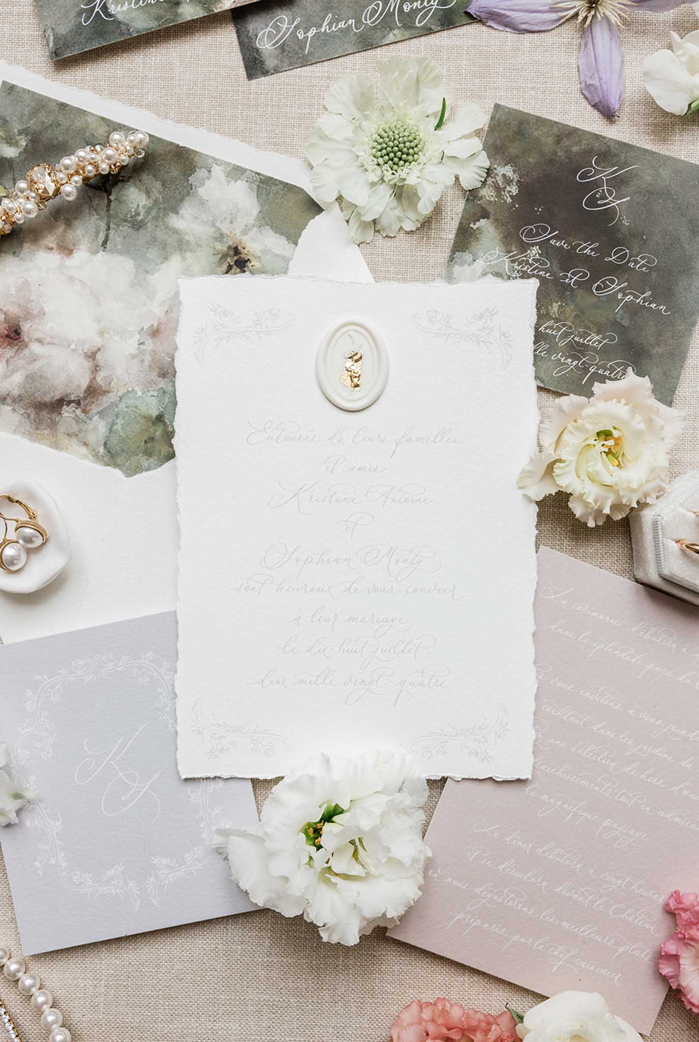 Flat lay of wedding stationery in sage and mauve tones with French calligraphy, scattered florals and gold jewelry