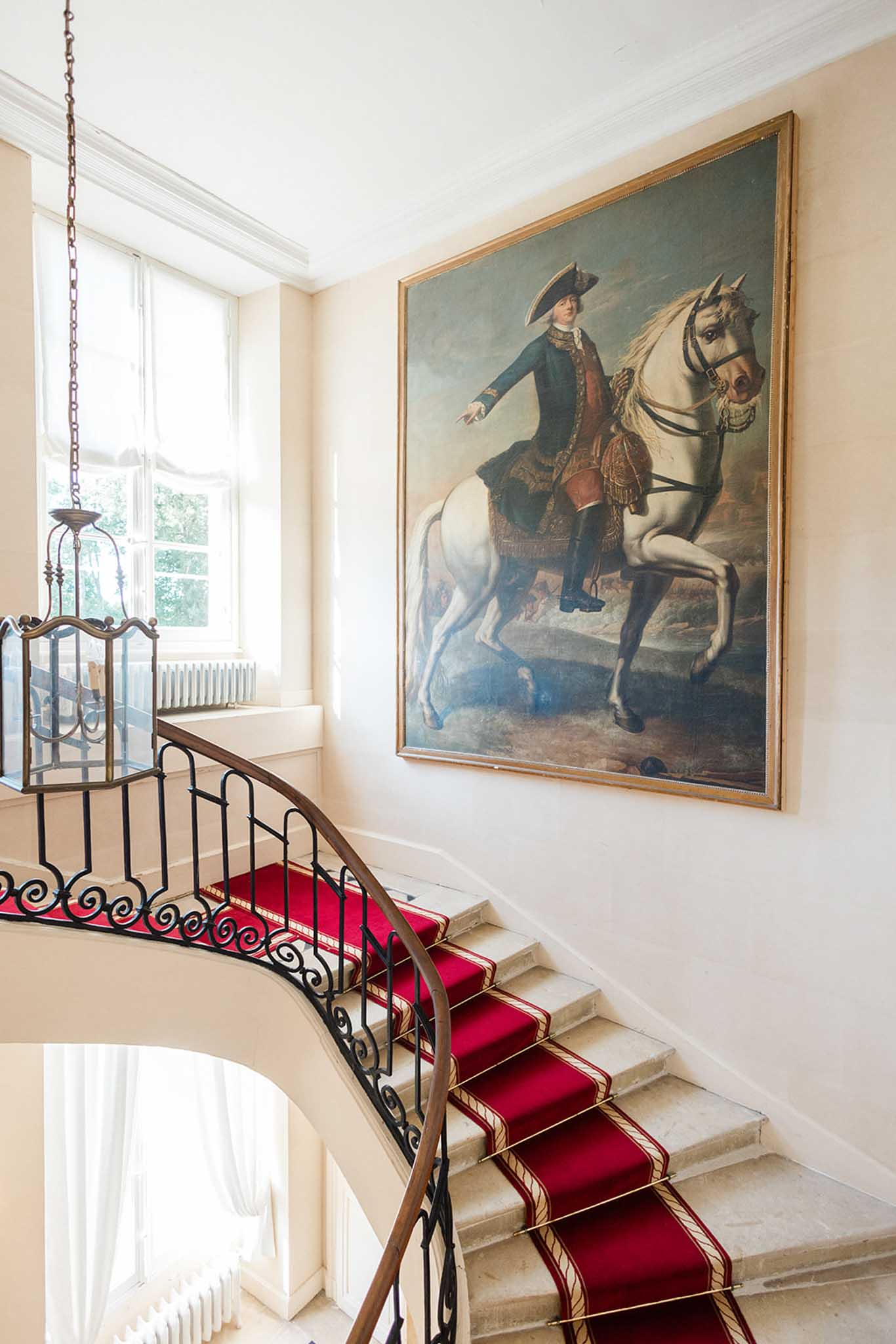 Grand staircase with wrought-iron railing, red carpet, and classical portrait in chateau interior