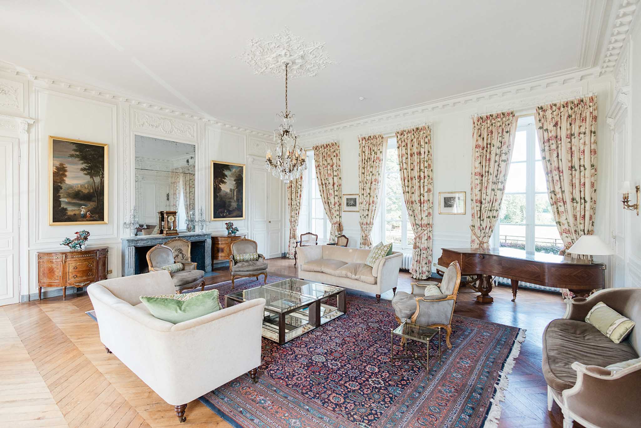 A wide-angle interior shot of a formal salon inside what appears to be a French château, with no wedding party or guests present. The room features white boiserie paneling with ornate plasterwork moldings and a decorative ceiling medallion from which a crystal and brass chandelier hangs. Furnishings include cream-upholstered sofas with sage green velvet cushions, Louis XVI-style gilt armchairs in grey velvet, and a glass-topped mirrored coffee table, all arranged over a large burgundy and navy Persian-style rug on herringbone parquet flooring. A dark marble fireplace with a gilt-framed mirror above it anchors one wall, flanked by large oil paintings in gold frames, while tall French windows dressed in floral pink and cream curtains line the opposite side of the room, with a walnut grand piano positioned beside them. The overall décor palette is cream, gold, and muted rose with classic French interior styling. Potential venue feature image.