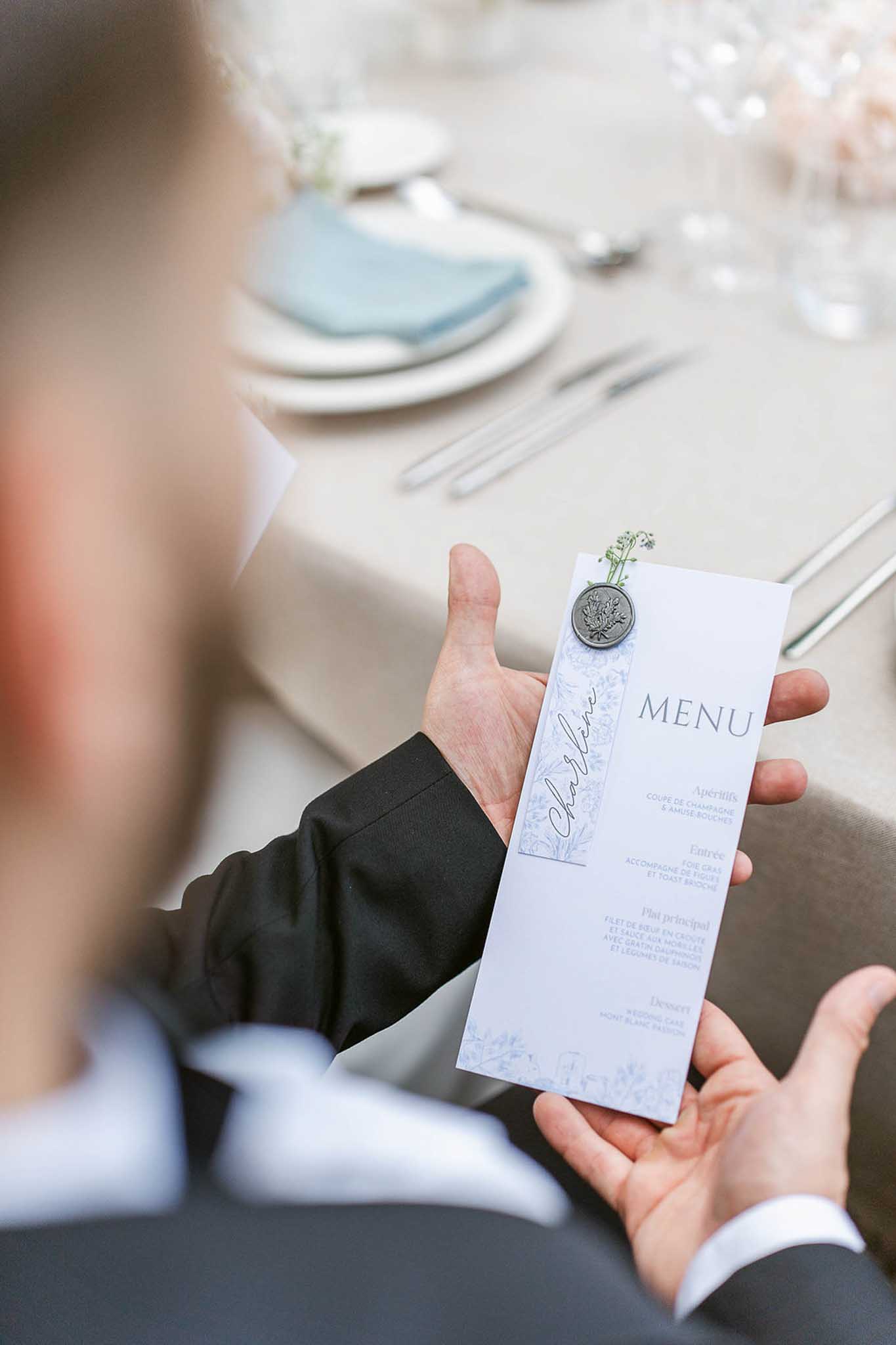 Groom's hand holding white menu card with wax seal and baby's breath sprig, formal place setting with blue napkin