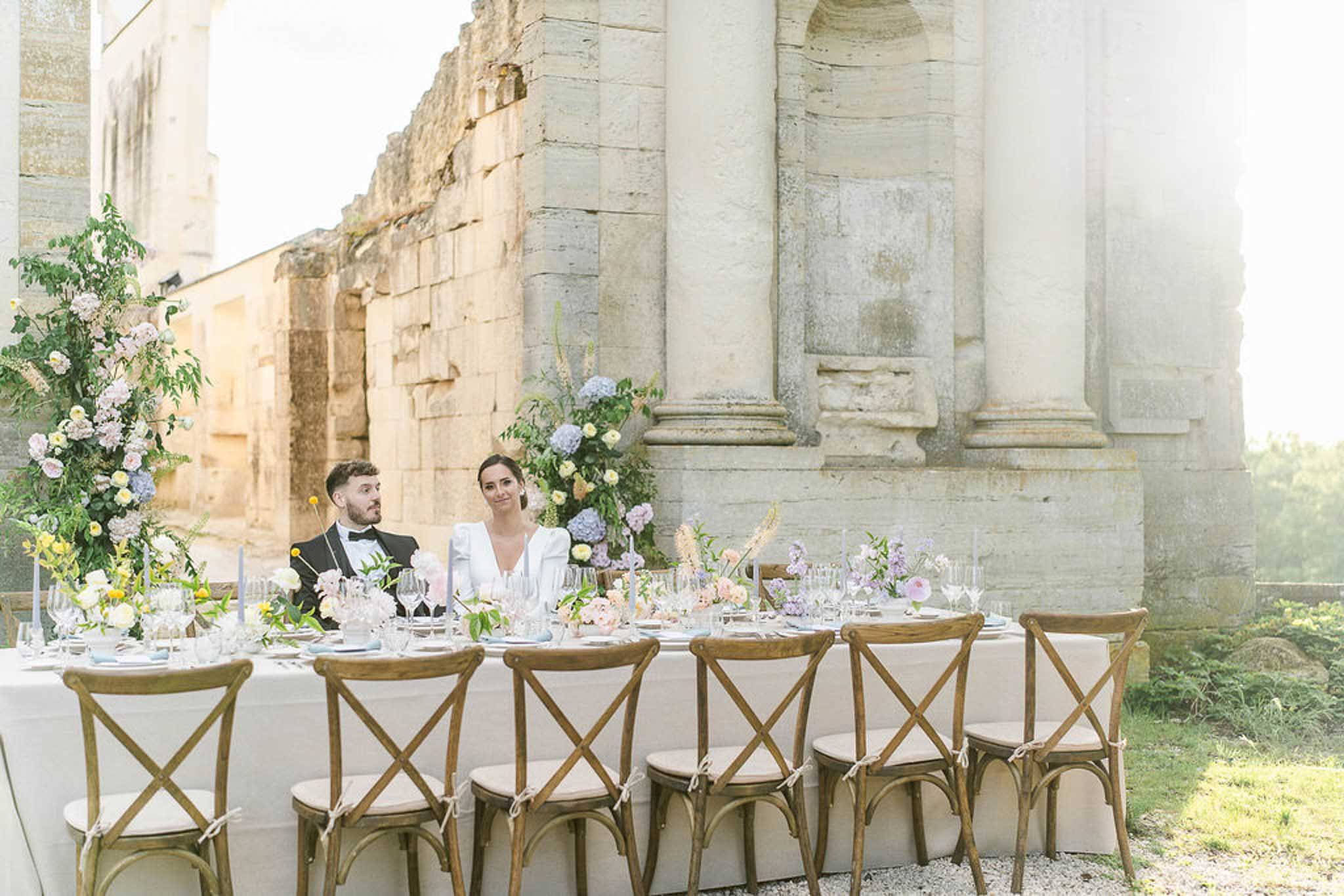 Bride and groom seated at outdoor reception table against historic stone columns with pink and lavender floral arrangements
