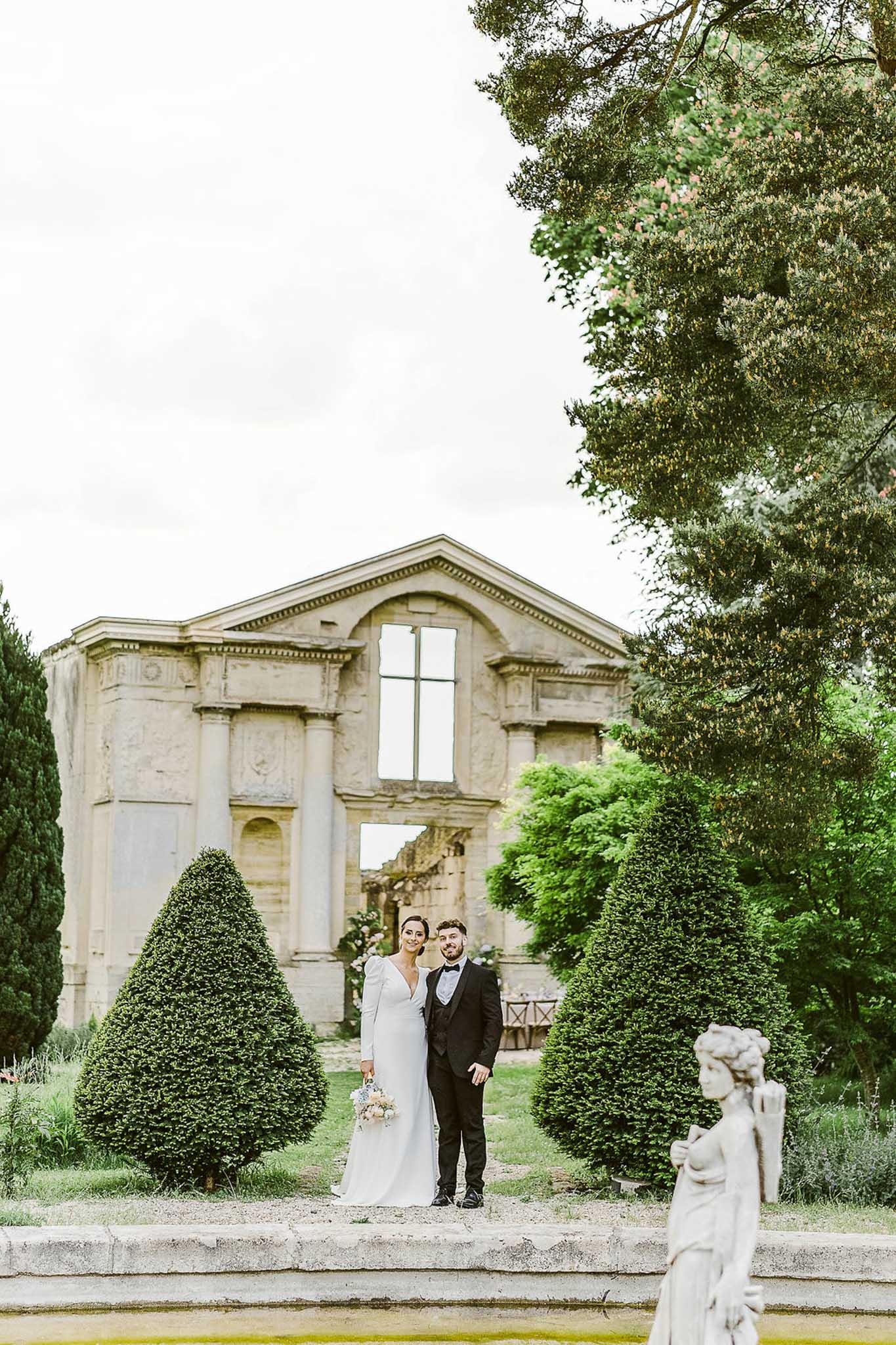 Bride and groom on gravel path between cone topiary with stone chapel and neoclassical gatehouse backdrop