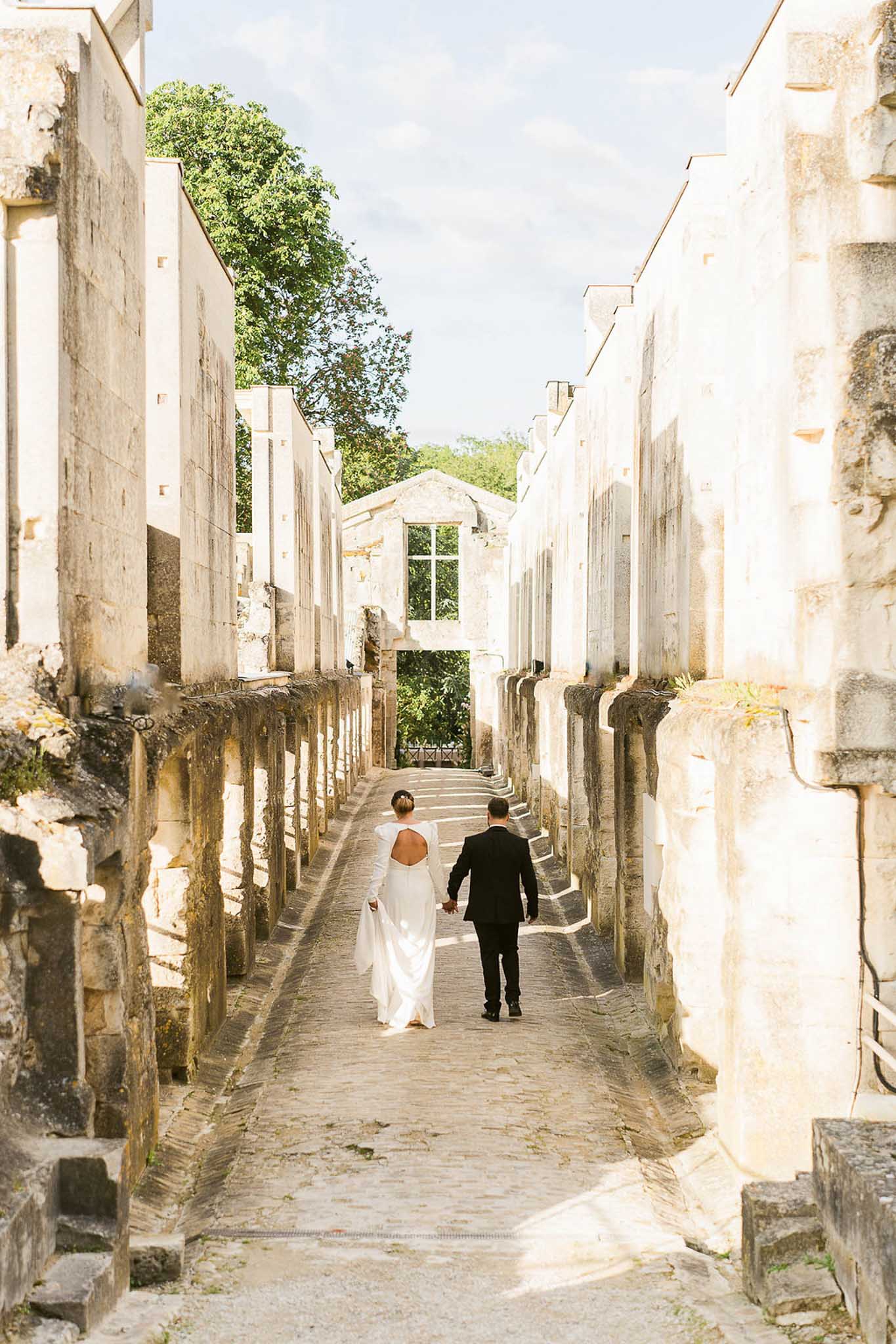 A couple portrait shot from behind showing the bride and groom walking hand-in-hand along a cobblestone path flanked by tall limestone ruins, likely an abandoned abbey or historic stone structure. The bride wears a fitted white long-sleeve gown with a deep open back and a short train, while the groom is dressed in a dark navy or black suit. The composition is a wide shot with strong leading lines created by the parallel stone walls drawing the eye toward an arched stone window frame and steps at the end of the passage. The setting has a raw, architectural quality with weathered pale limestone walls and dappled sunlight casting sharp shadows across the path. Potential venue feature image.