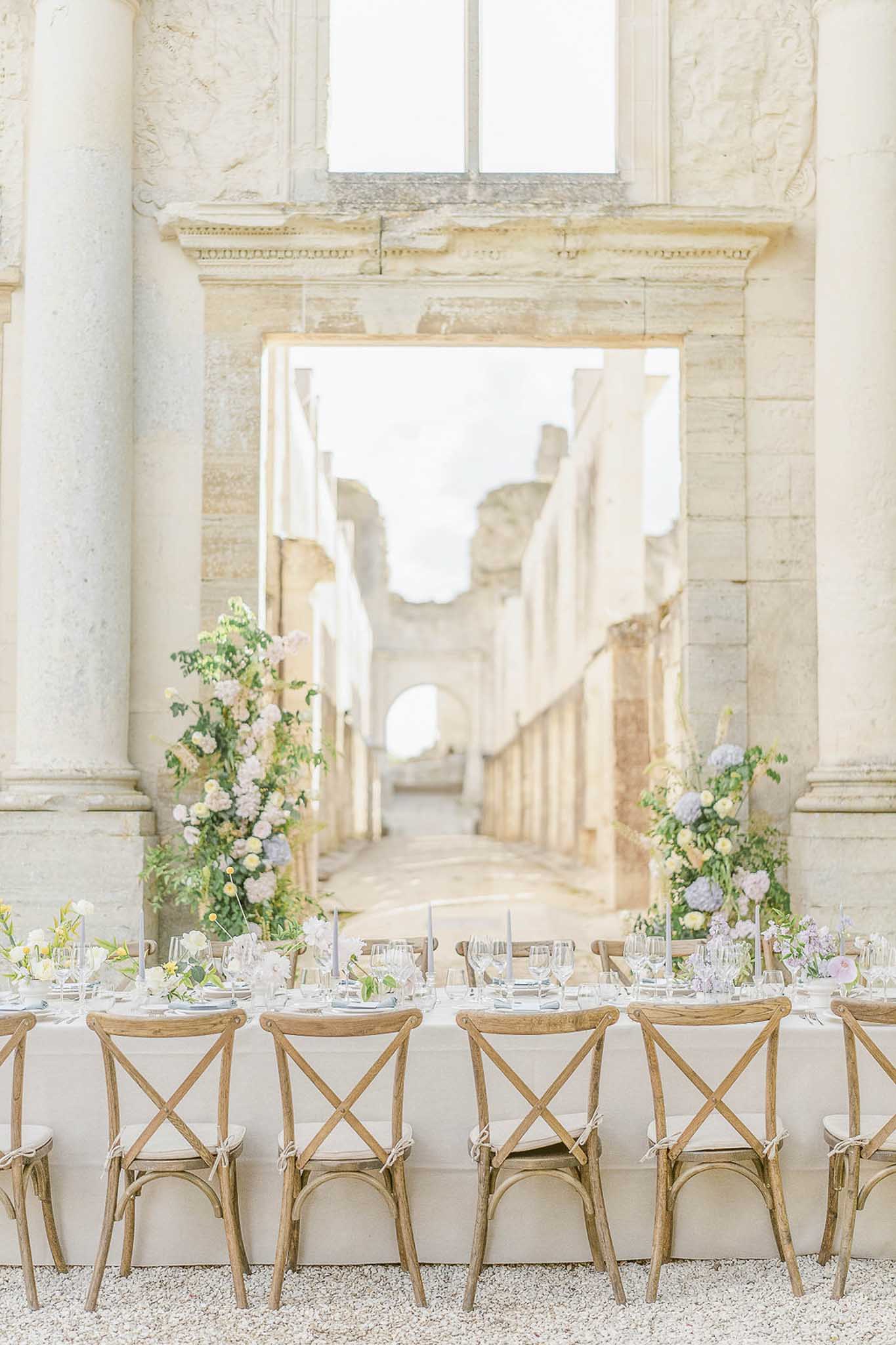Long reception table with white linen and pink-blue floral arrangements in arched stone courtyard