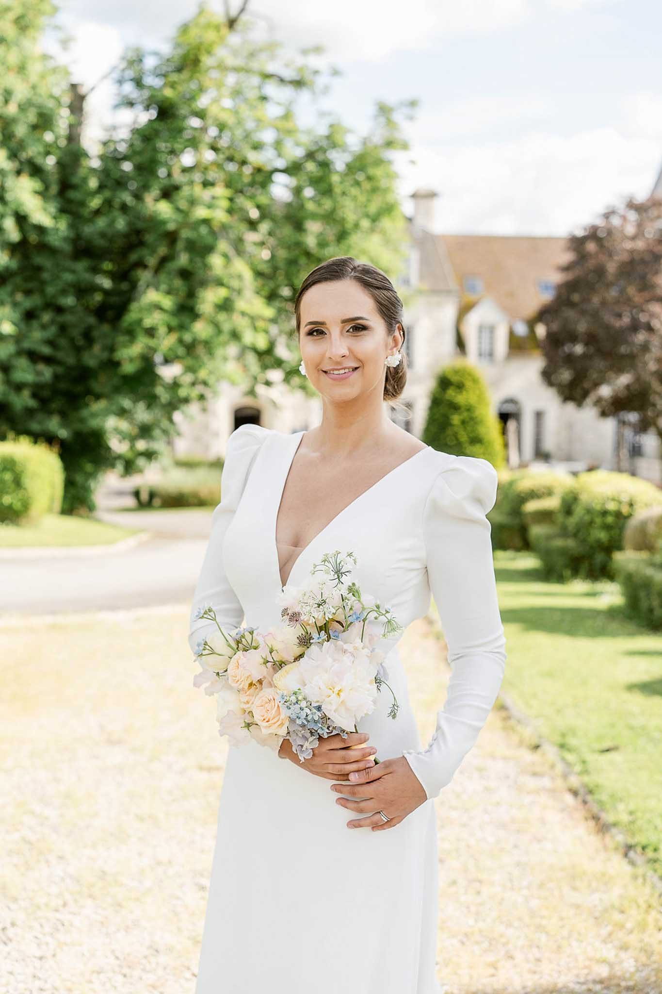 Bride holding bouquet of cream roses and dusty blue hydrangeas in chateau courtyard