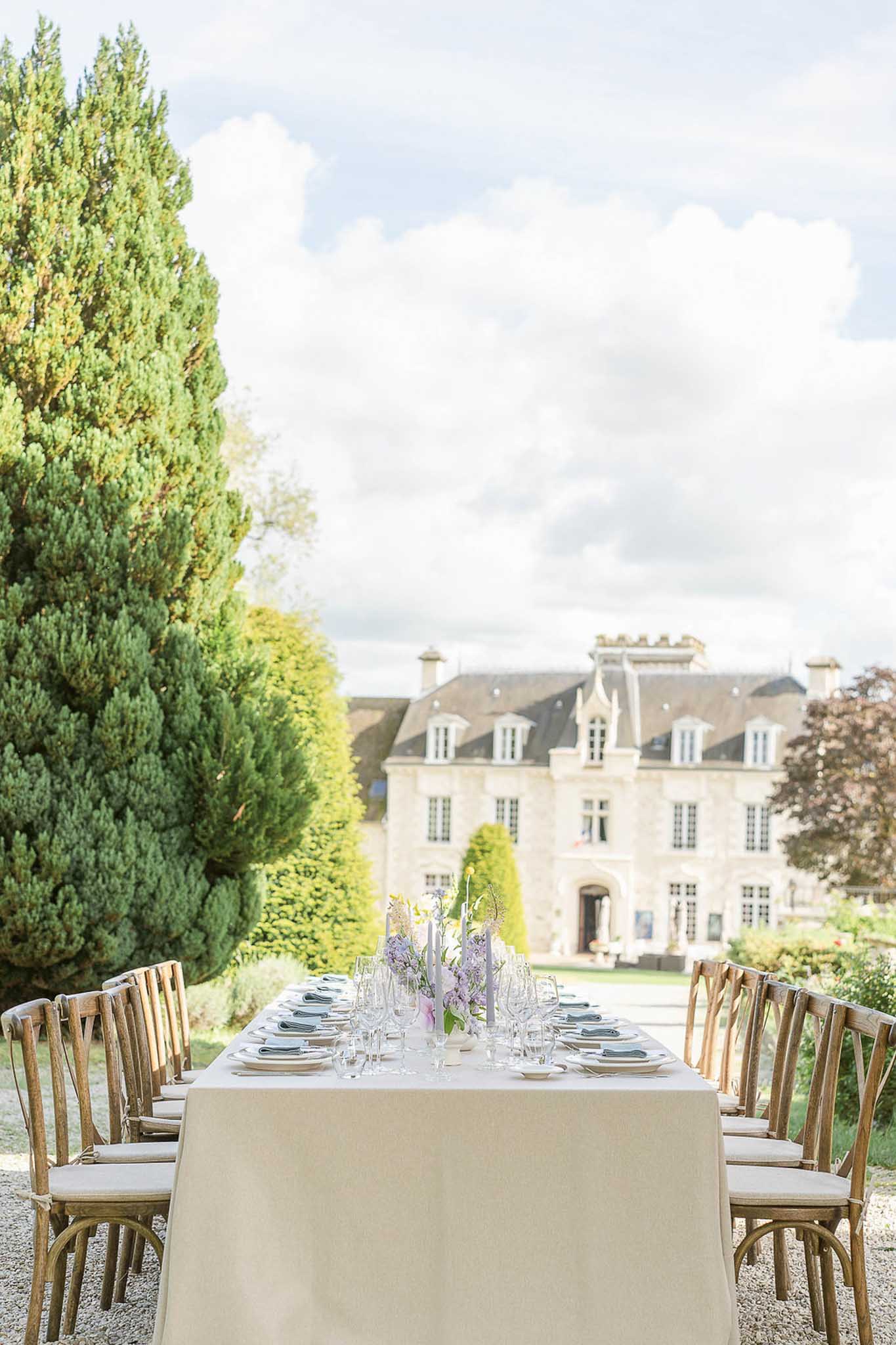 Outdoor reception table toward classical chateau, cream linen, cypress hedges, lavender floral centerpiece center