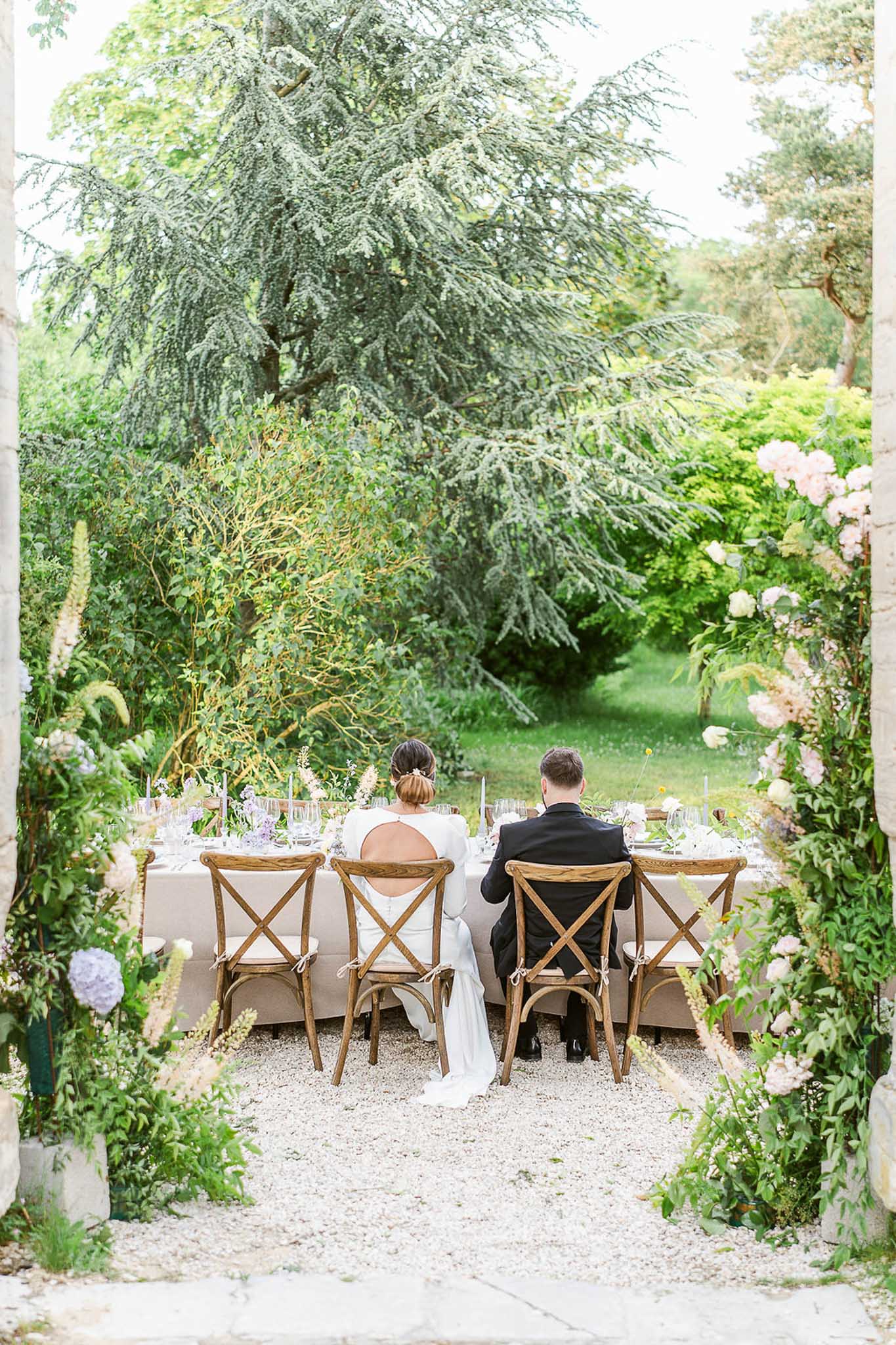 Bride and groom at head of outdoor reception table beneath flowering garden archway at a French estate