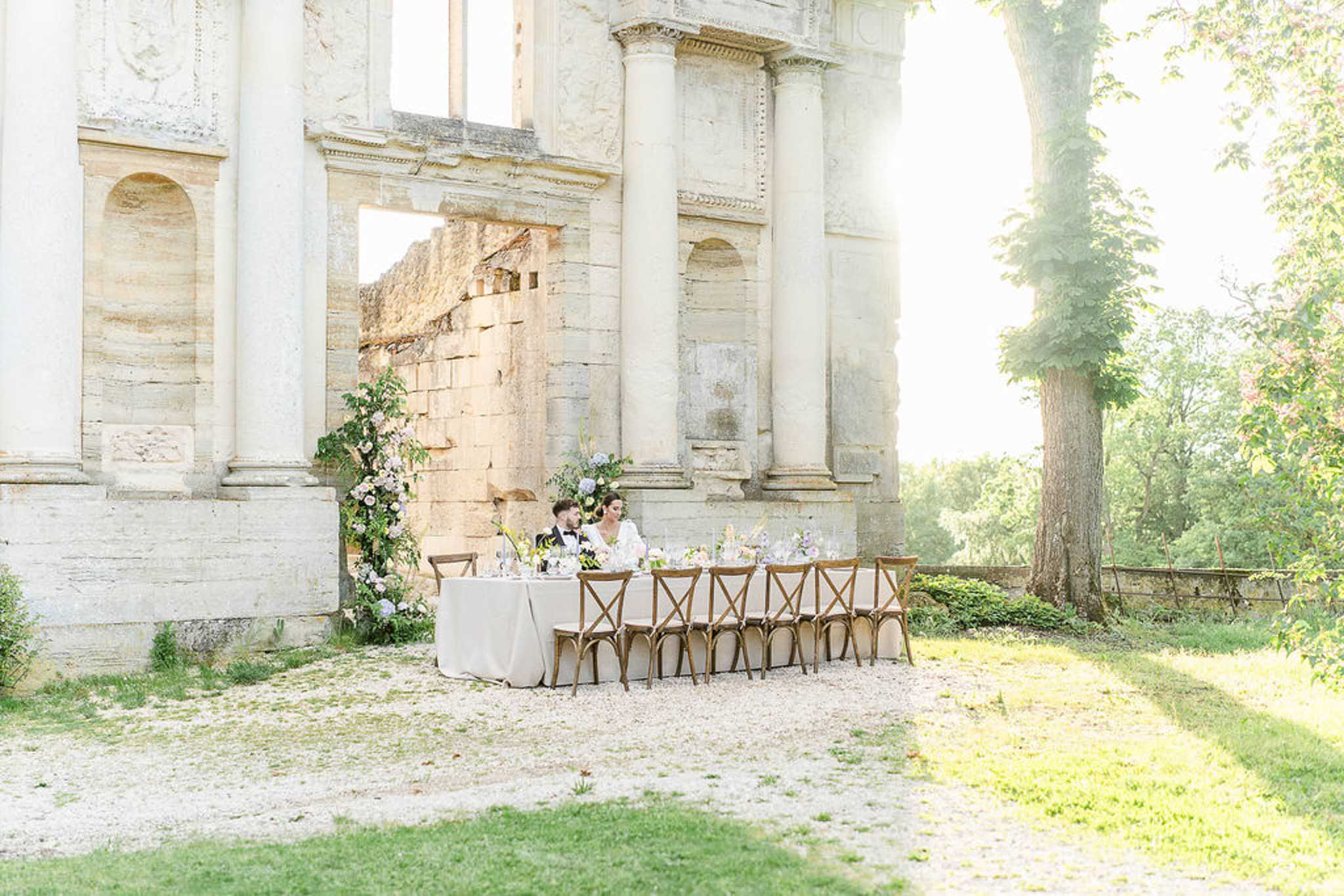 Couple at a sweetheart table beneath a floral arch against a backdrop of classical stone ruins with columns in a historic courtyard.