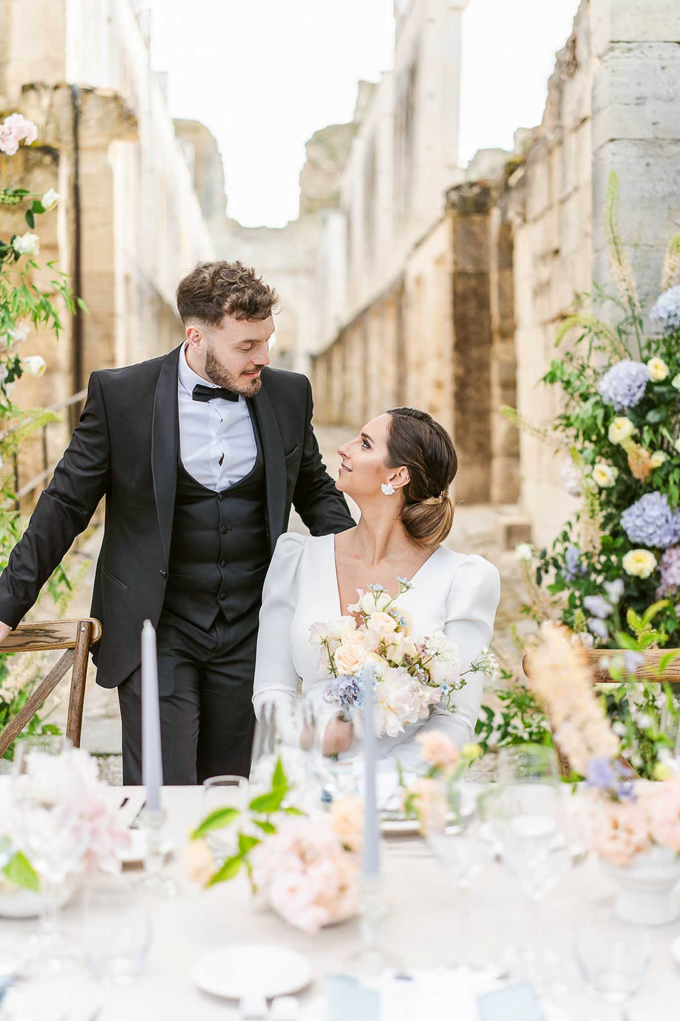 Bride in ivory long-sleeve dress with peach-pink bouquet and groom in black tuxedo walk through floral-arch reception corridor