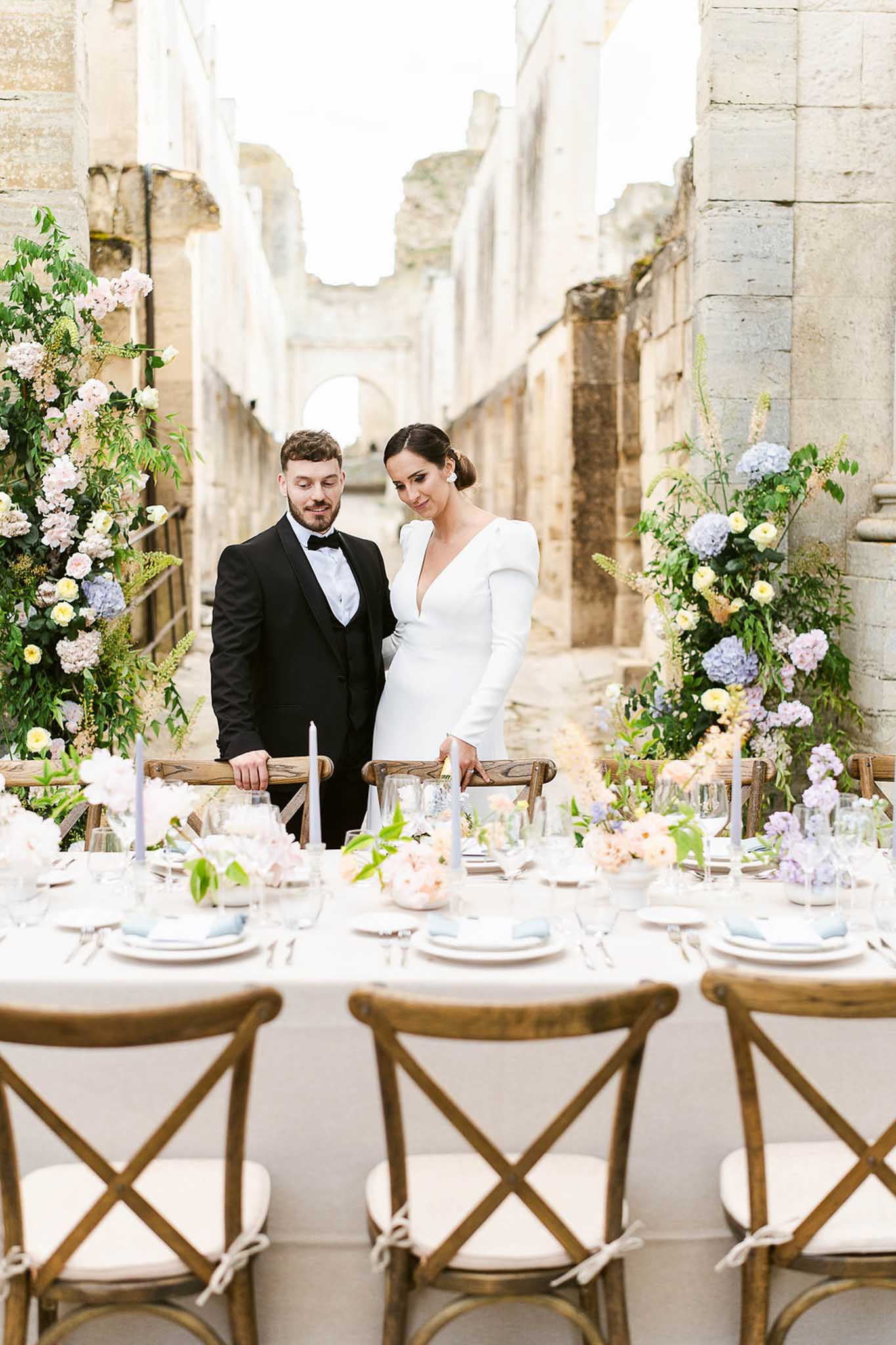 Bride and groom at outdoor banquet table in stone courtyard with tall floral arrangements of pink roses and blue hydrangeas