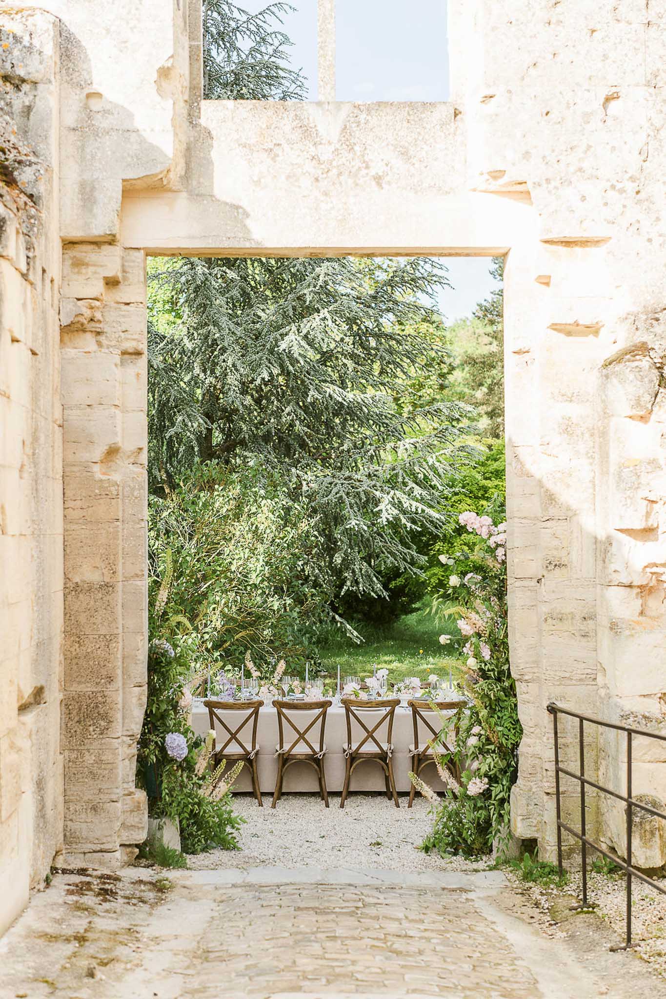 Outdoor reception table framed through a stone archway with pink and lavender floral centerpieces and wooden chairs