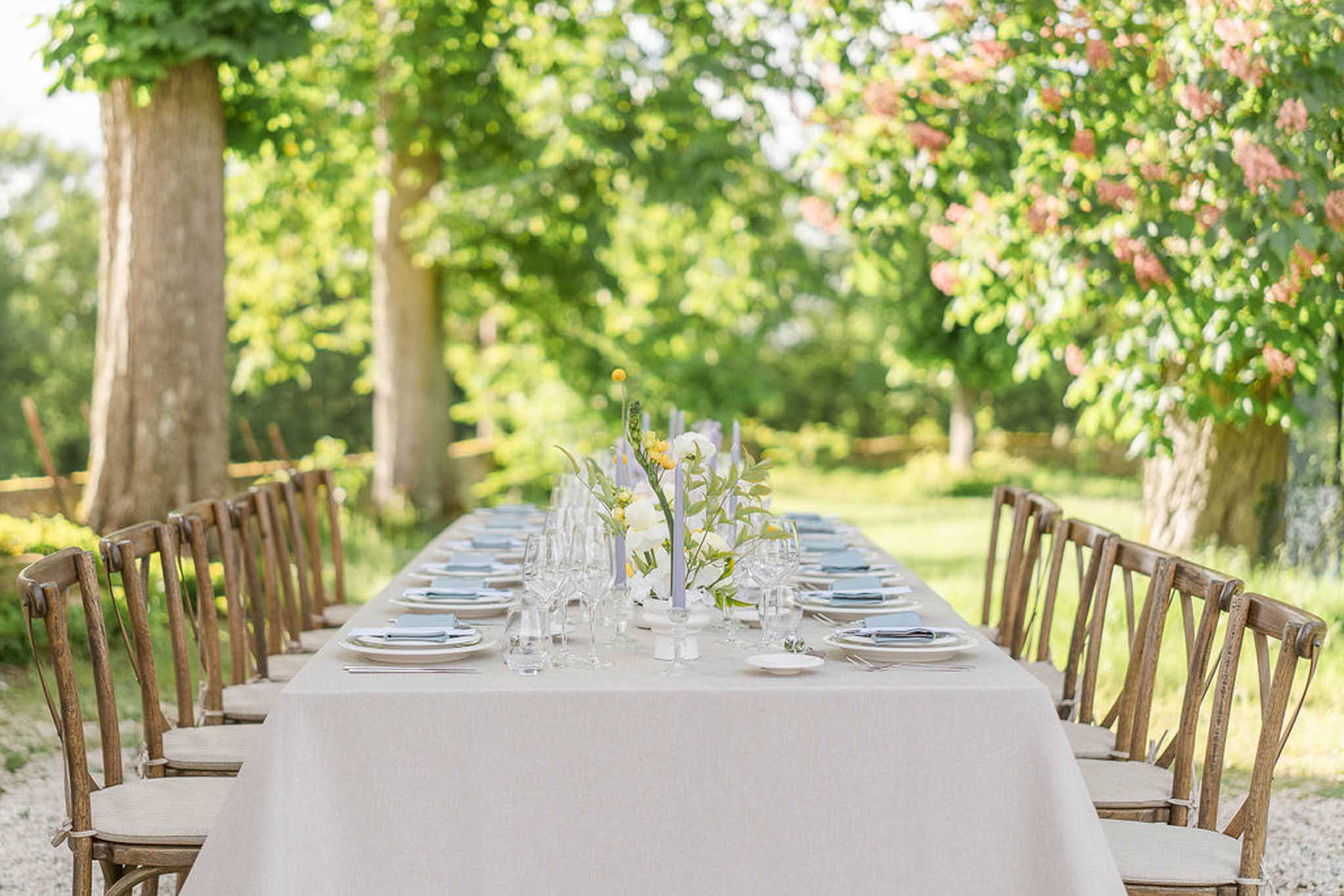 Long outdoor reception table with ivory linens and slate-blue napkins set beneath tree canopy in garden